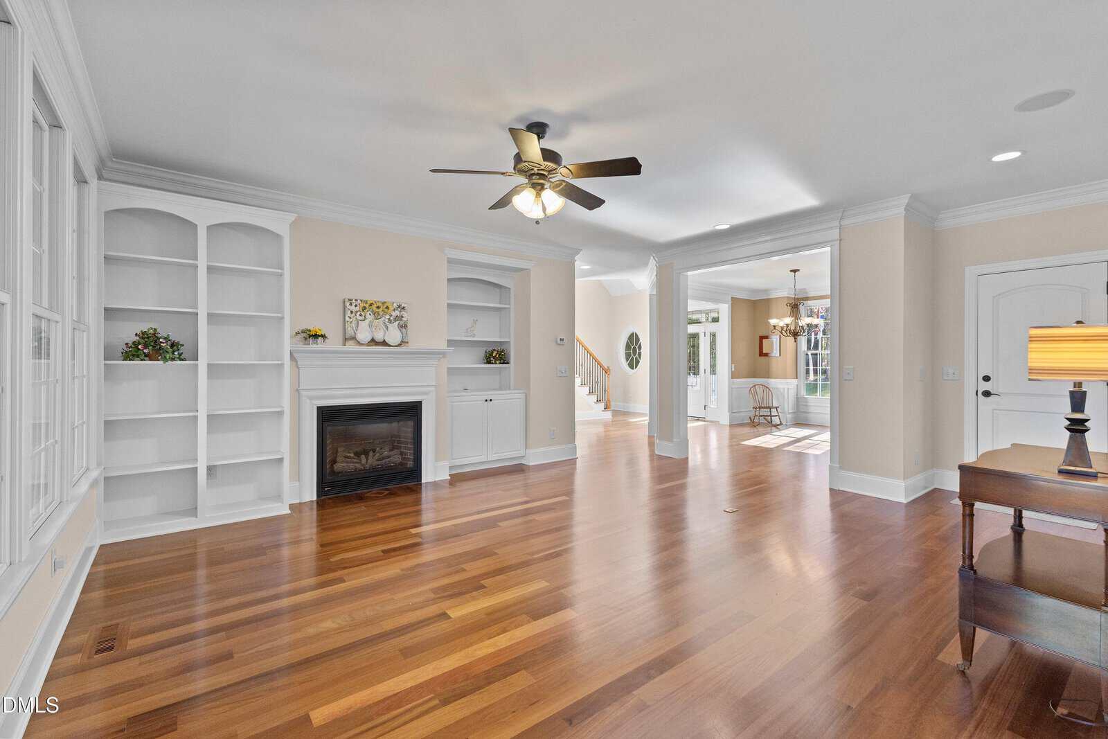 Bright living room with hardwood floors, built-in white shelves, a fireplace, ceiling fan, and large windows. The space opens to a hallway and other rooms with natural light streaming in.