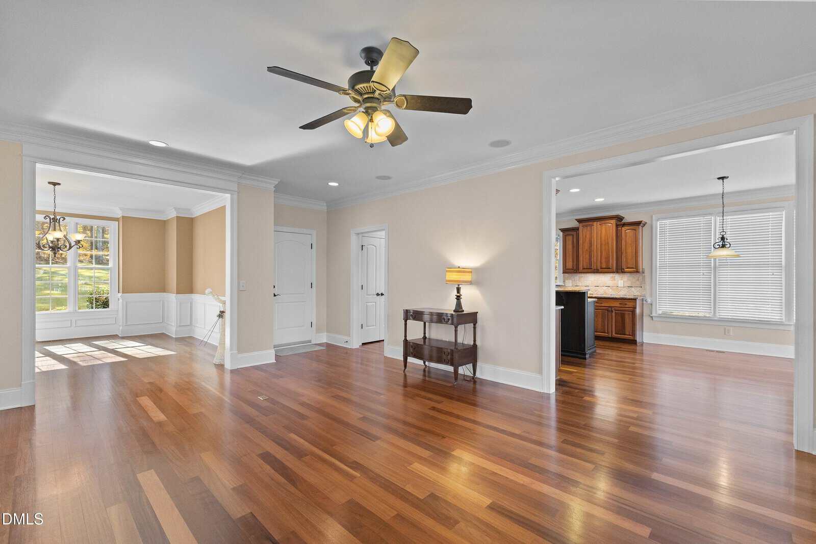 Spacious living area with hardwood floors, beige walls, ceiling fan, wall lamp, and wood furniture, leading to a sunlit dining room with large windows and a kitchen with wooden cabinets.