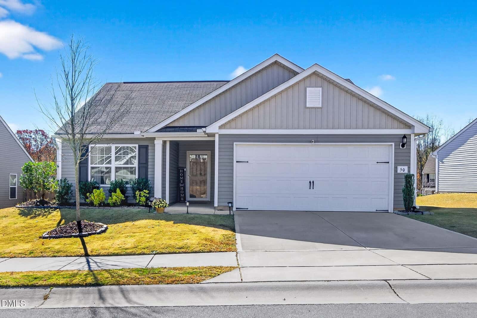 Single-story suburban house with gray siding, white trim, and a double garage located at 50 Shallow Drive in Youngsville. There is a small front porch, minimal landscaping, and a driveway leading to the street. The sky is clear and blue.