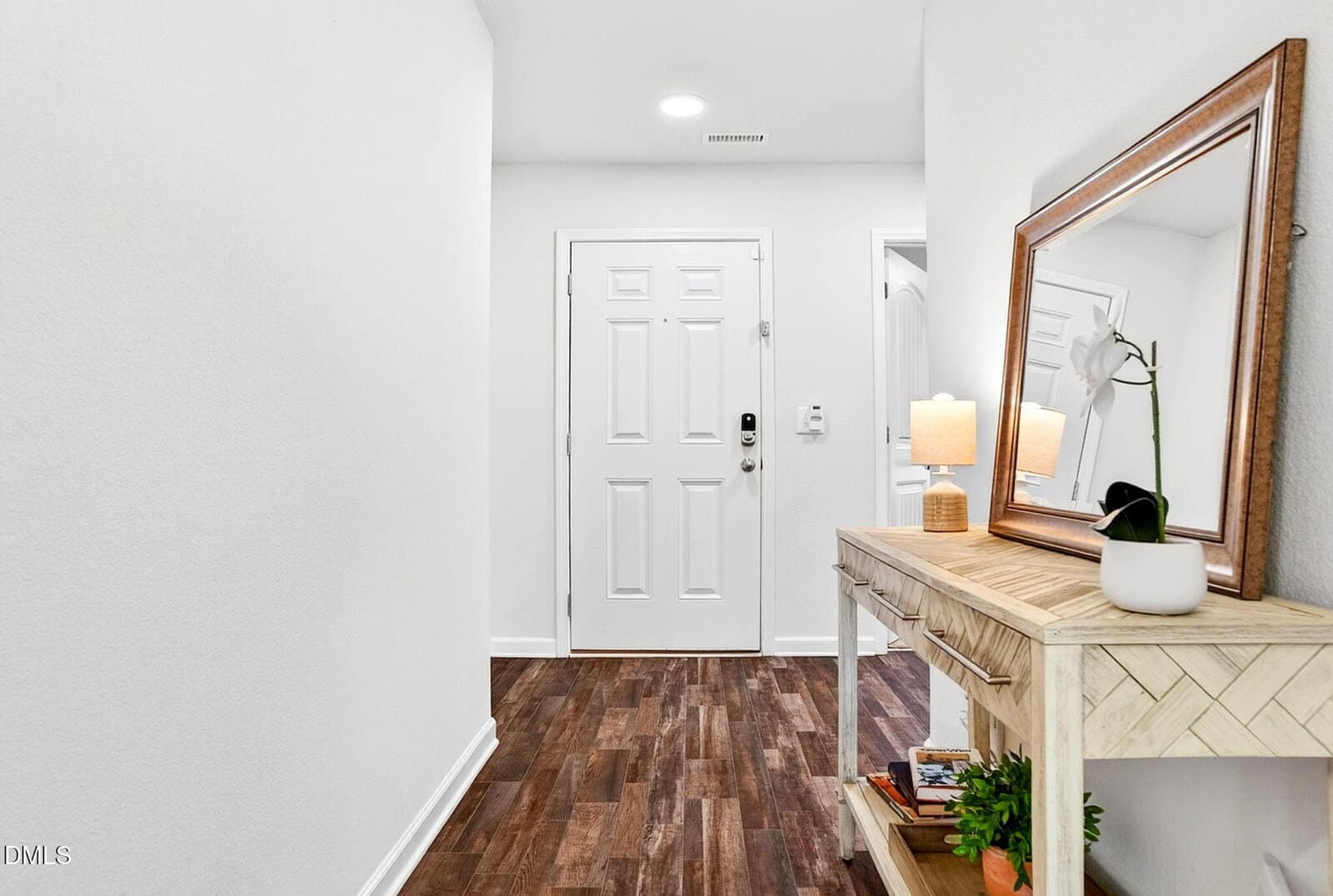 A hallway with wood flooring leads to a white front door. There is a light-colored console table with a lamp, a potted plant, books, and a framed mirror on top. The walls are plain white.