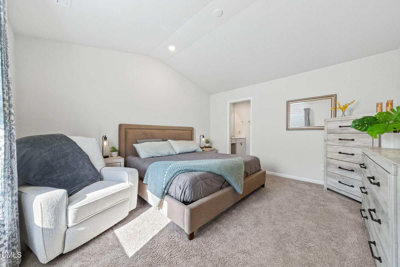 A bedroom with a beige upholstered bed, blue blanket, white armchair, gray dresser with decor, and carpeted floor. Natural light enters through a window; a door leads to an adjoining bathroom. The walls are painted white.