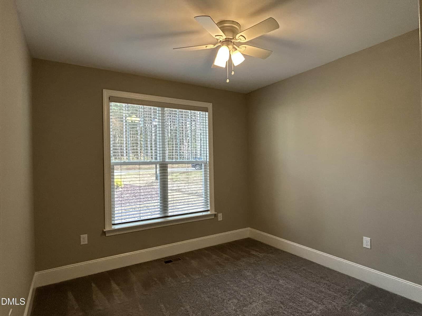 Empty room with beige walls, white trim, a large window with blinds, a ceiling fan with lights, and carpeted floor. Natural light comes through the window, revealing a view of trees outside.