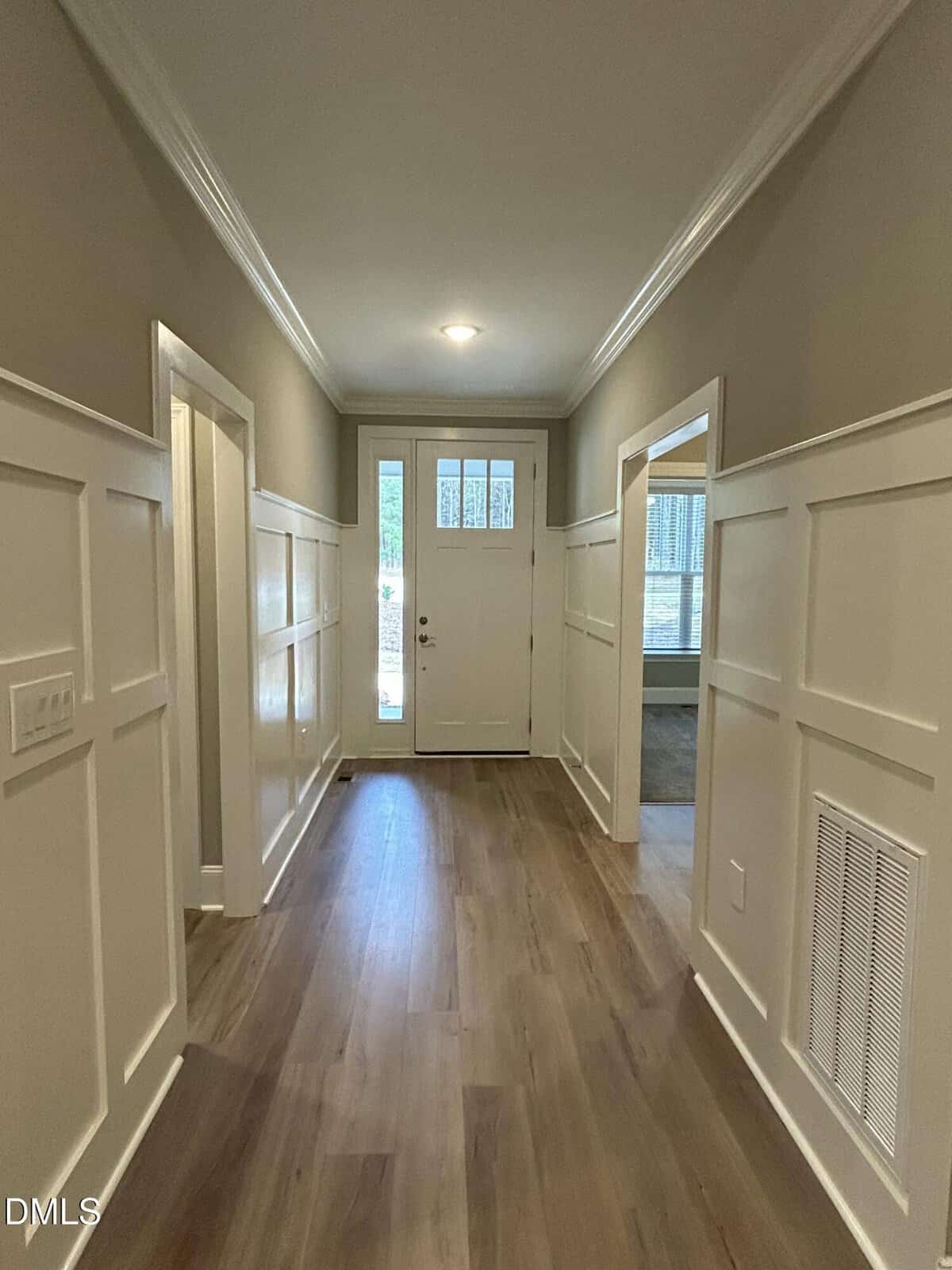 A hallway with wood flooring, white wainscoting, beige upper walls, and a white front door with glass panels. There are several doorways on both sides and a ceiling light at the far end.