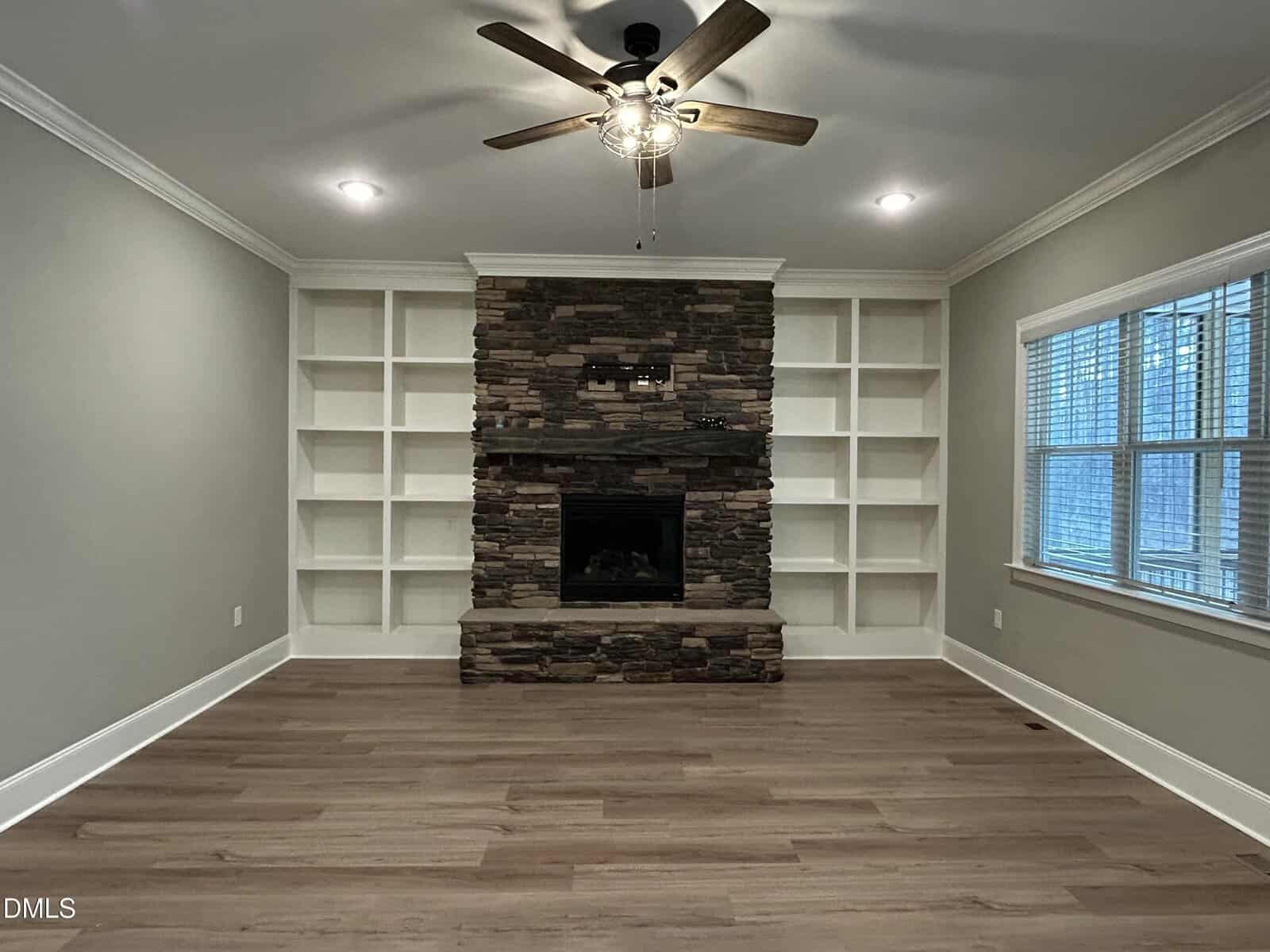A living room features a stone fireplace with built-in white shelves on both sides, wood plank flooring, neutral walls, a ceiling fan with lights, and a large window with blinds on the right wall.