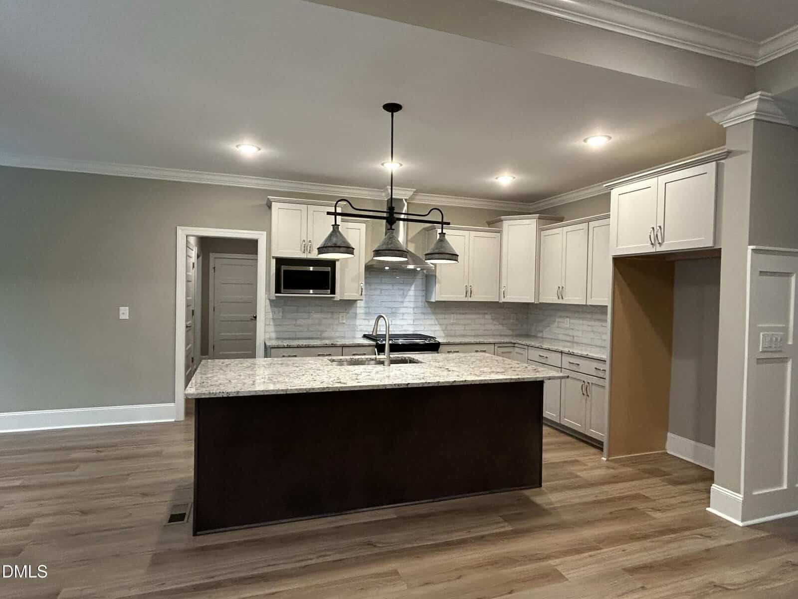 A modern kitchen with white cabinets, a large center island with a sink, stainless steel appliances, subway tile backsplash, wood flooring, and overhead lighting fixtures. The walls are painted gray.