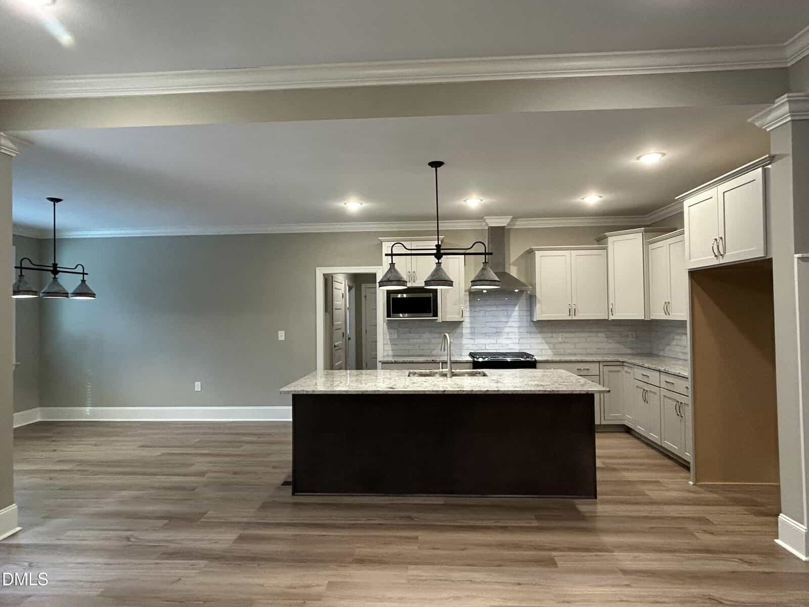 Modern kitchen with a large island featuring a marble countertop, white cabinets, stainless steel appliances, pendant lighting above the island, and light wood flooring. An adjacent dining area is visible to the left.