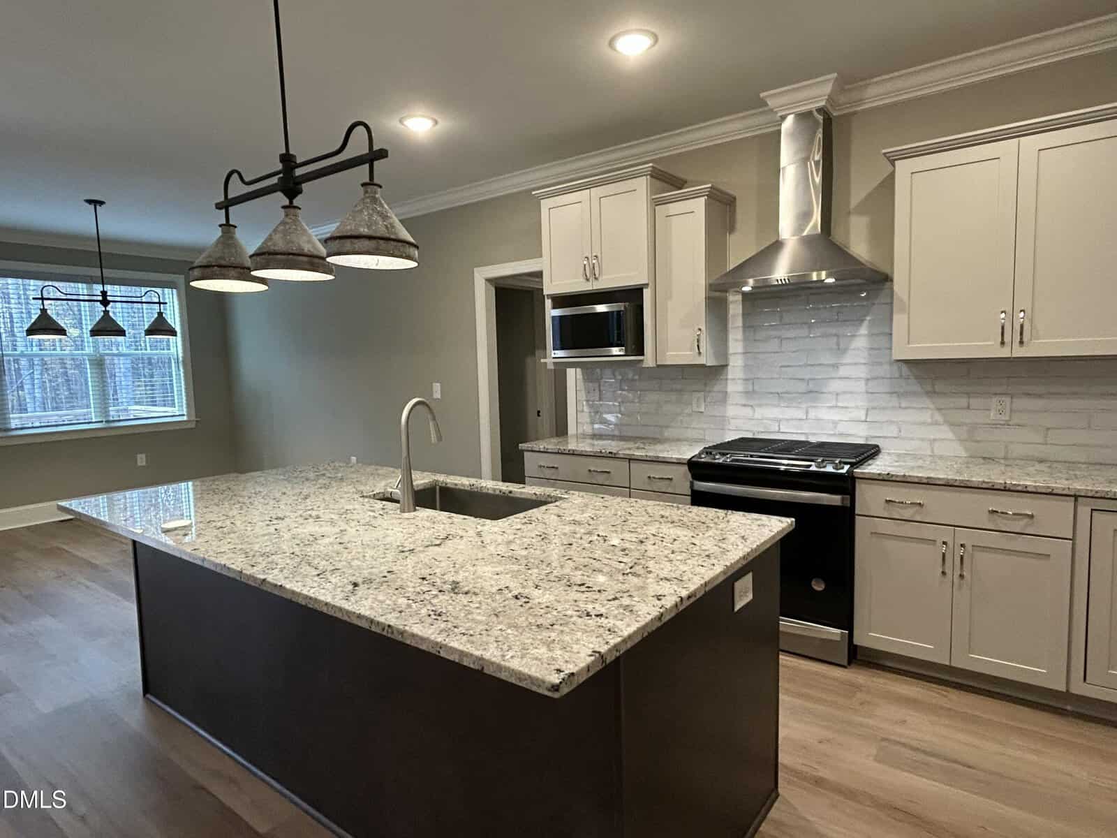 Modern kitchen with a large granite island, farmhouse-style pendant lights, stainless steel appliances, light gray cabinets, white subway tile backsplash, and wood flooring. Dining area with large windows in the background.