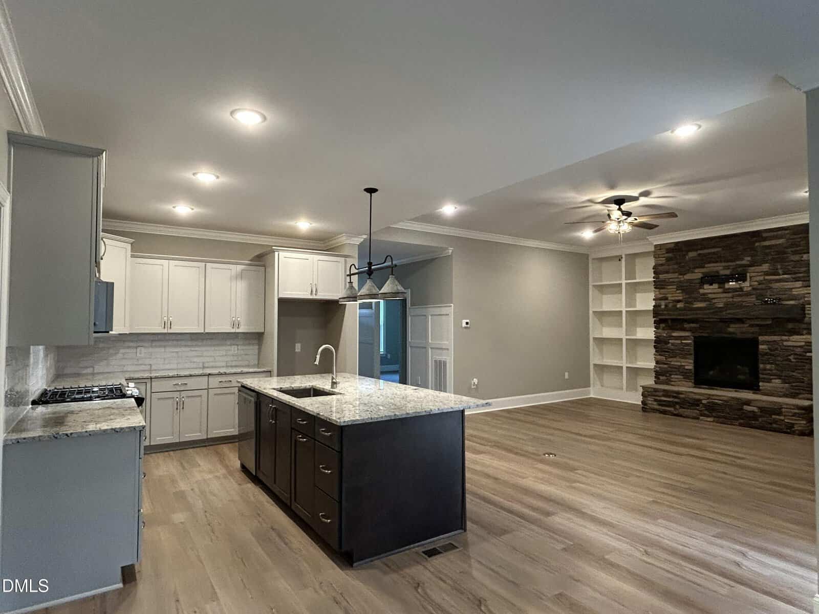 Open-concept kitchen and living area with light wood flooring, white cabinets, a central island with dark cabinetry, built-in shelves, a stacked stone fireplace, and recessed lighting. Ceiling fan is also present in the living area.