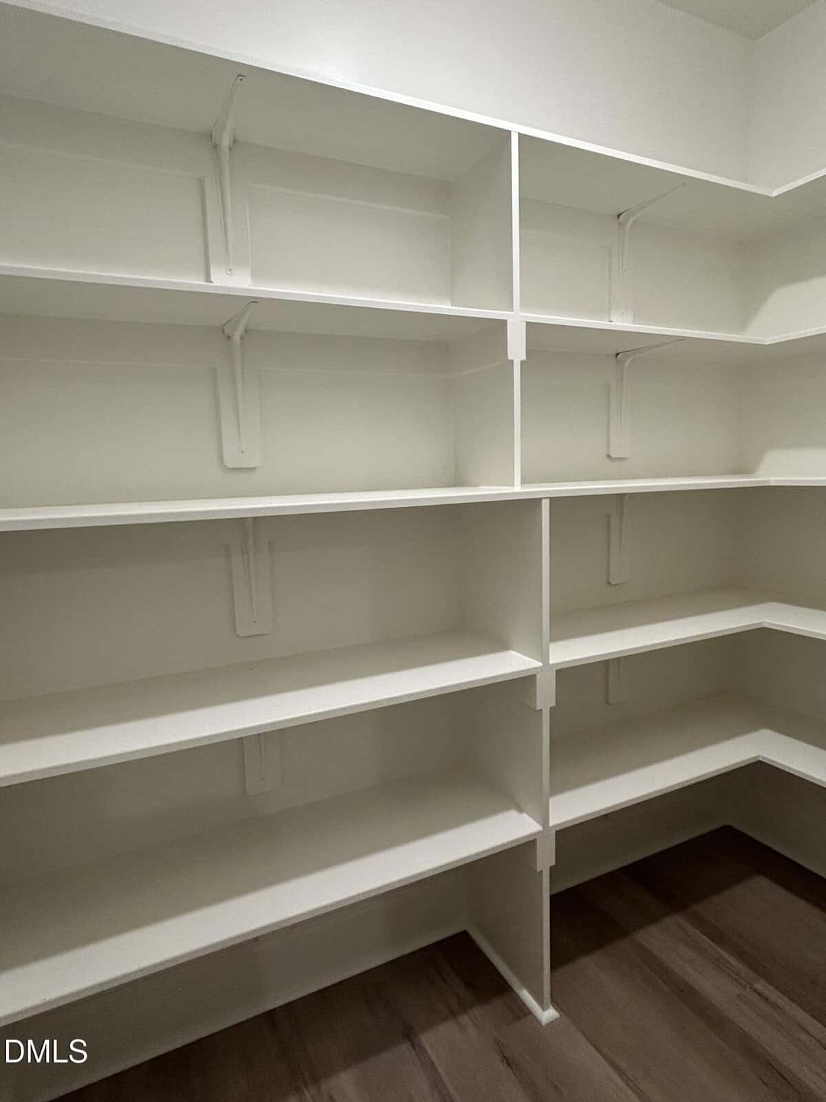 Empty white built-in shelves line the walls of a walk-in pantry with dark wood flooring. Shelves are supported by white metal brackets and extend around the corner, providing ample storage space.