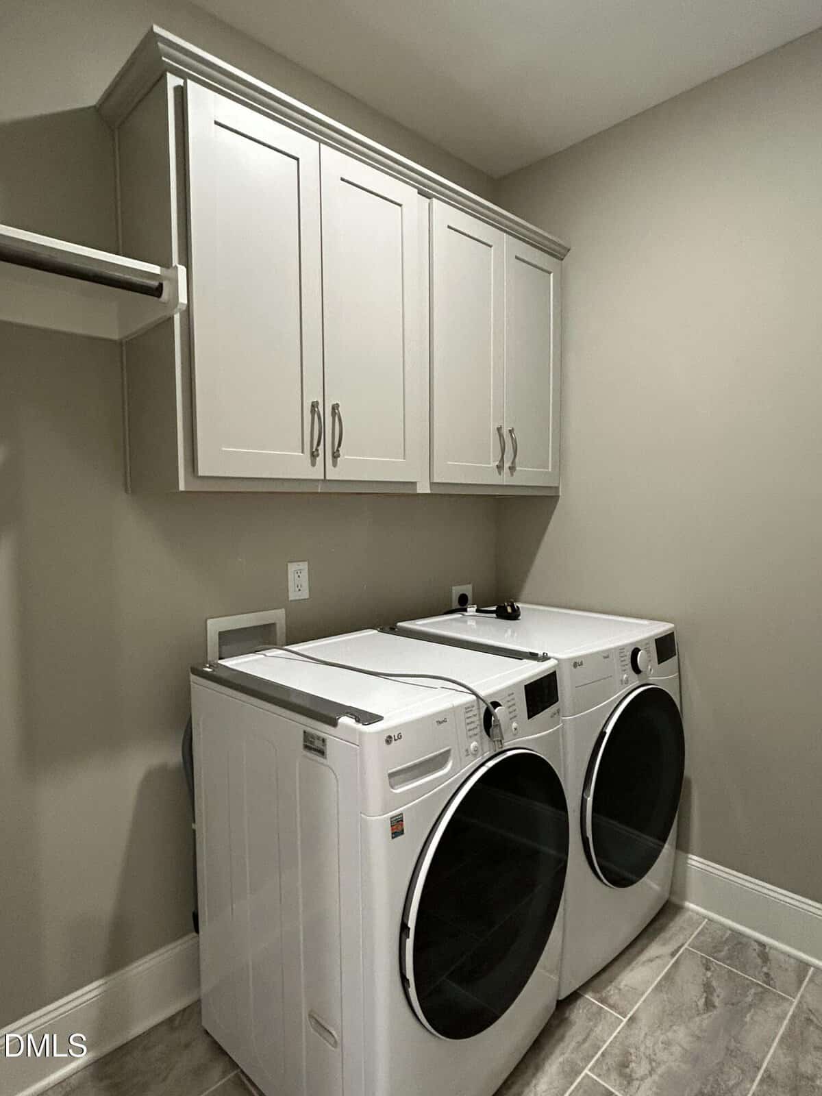 A laundry room with a front-loading washer and dryer side by side beneath white cabinets, beige walls, and tile flooring. An empty wall-mounted shelf is visible on the left.