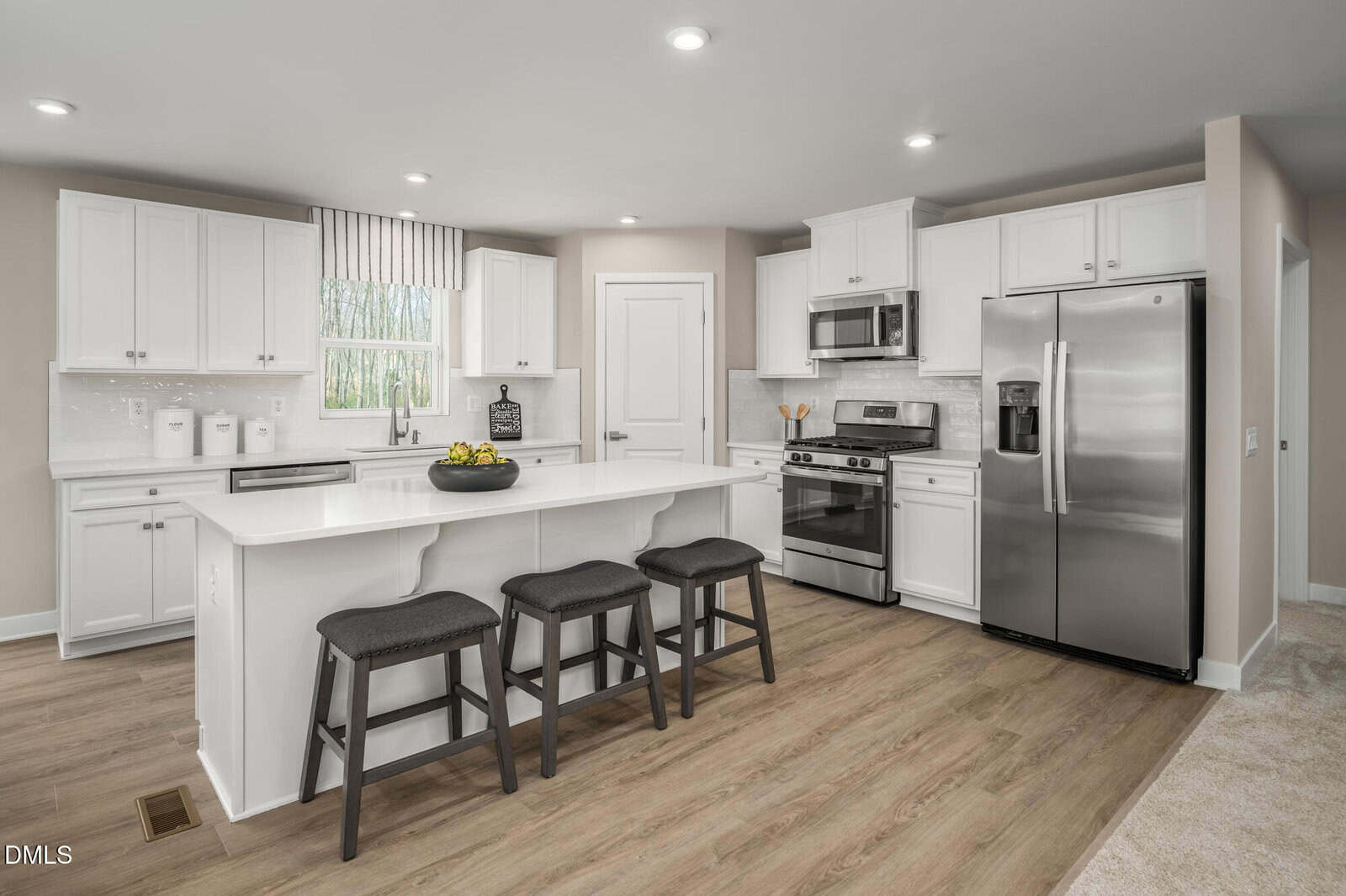 Modern kitchen with white cabinets, stainless steel appliances, a center island with three black stools, light wood flooring, and a window with striped curtains above the sink. A bowl of fruit sits on the island.