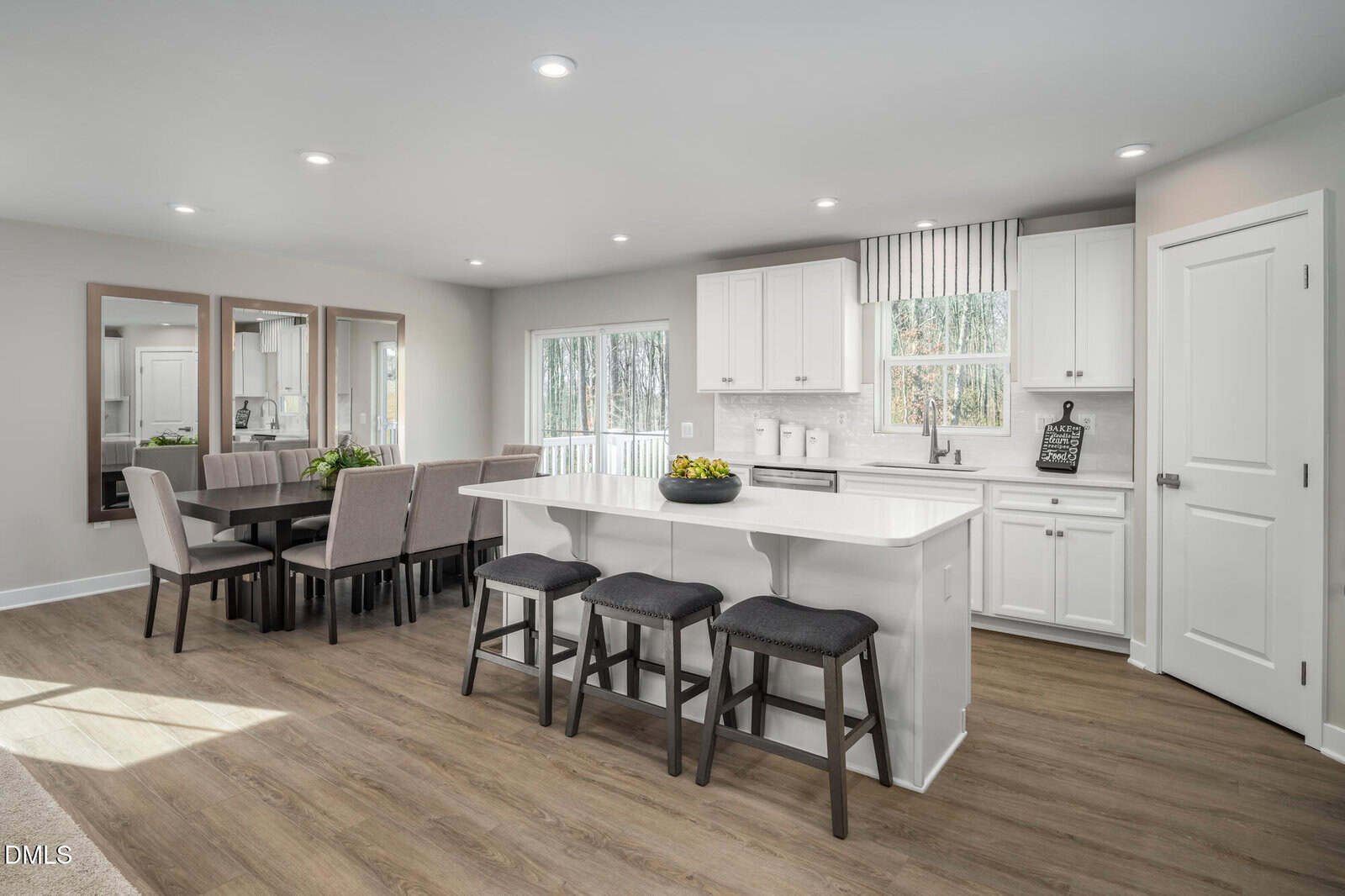 A modern kitchen with white cabinets, stainless steel appliances, and a large island with three dark stools. The adjoining dining area has a table with eight upholstered chairs and three mirrors on the wall. Wood flooring throughout.