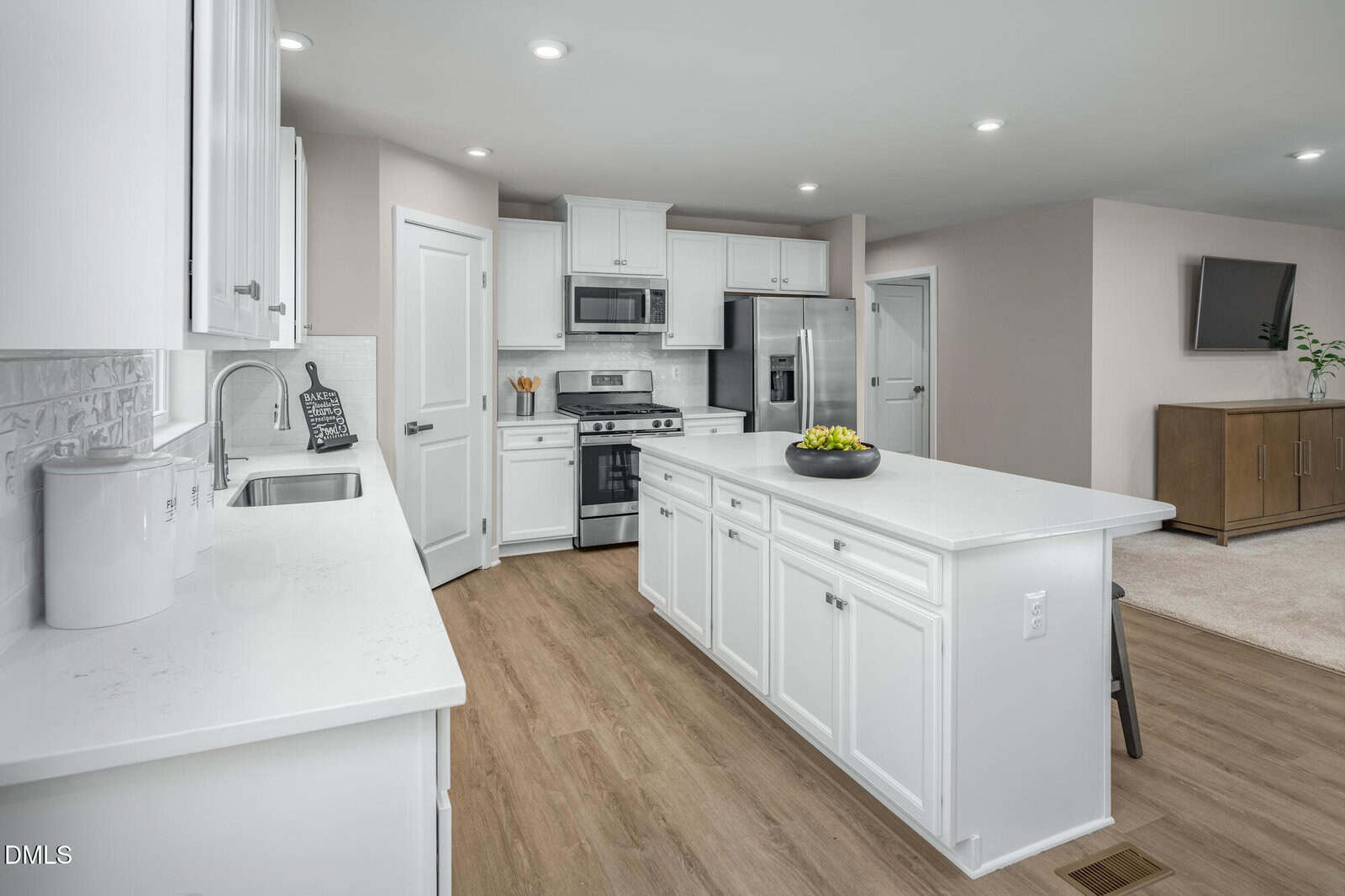 Modern kitchen with white cabinets, stainless steel appliances, and a large island with a bowl of apples. Light wood flooring and recessed ceiling lights. The kitchen opens to a living area with a TV and cabinet.