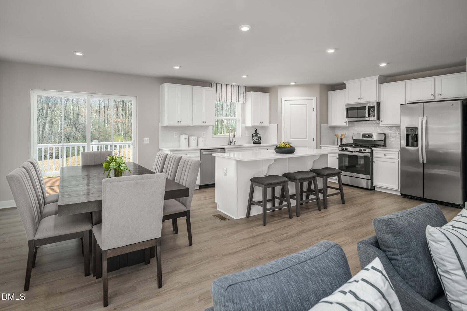 Modern kitchen and dining area with white cabinets, stainless steel appliances, a kitchen island with three stools, a large dining table with eight chairs, wood flooring, and a view of a deck and trees through glass doors.
