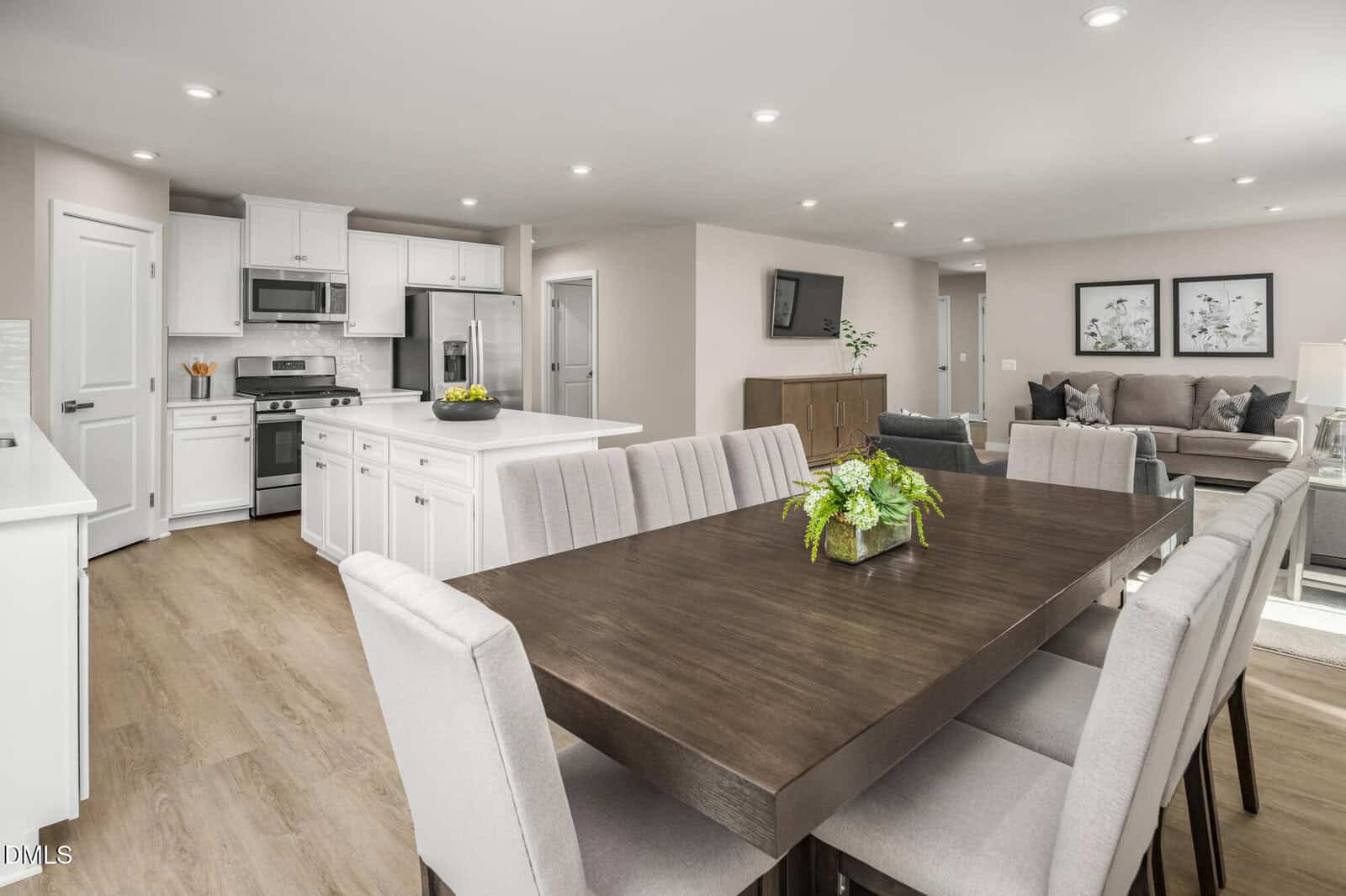 Open-concept space with a dining area, dark wood table, eight upholstered chairs, adjacent white kitchen with island, stainless steel appliances, and a living area with a sofa, chairs, and framed wall art.