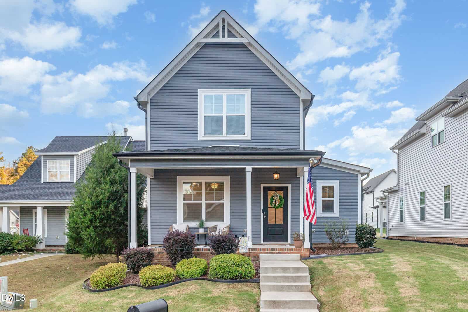 A two-story gray house with white trim at 203 Kipling Drive, Oxford, features a front porch with white railings, a wreath on the door, an American flag, and neatly trimmed shrubs. Steps lead from the sidewalk to the welcoming porch.