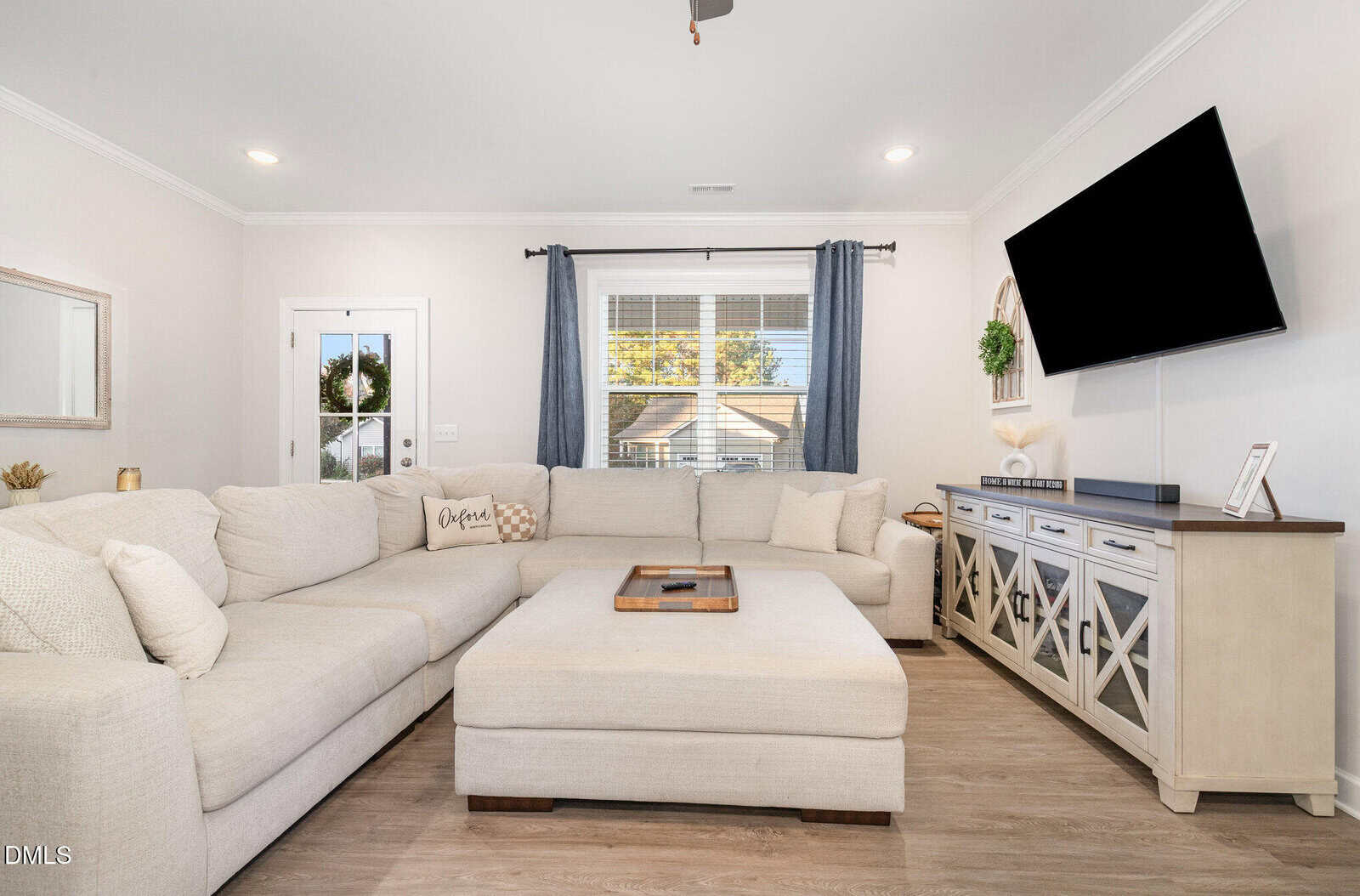 A bright living room with a large beige sectional sofa, matching ottoman, light wood floors, a wall-mounted TV, and a console with decorative items. A window with gray curtains lets in daylight.