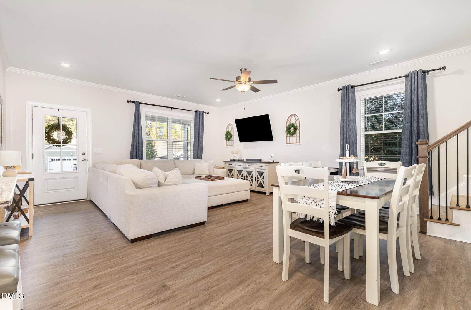 A modern open-concept living and dining area with light wood flooring, beige sectional sofa, wall-mounted TV, white dining table with six chairs, and large windows with gray curtains. A staircase is visible on the right.