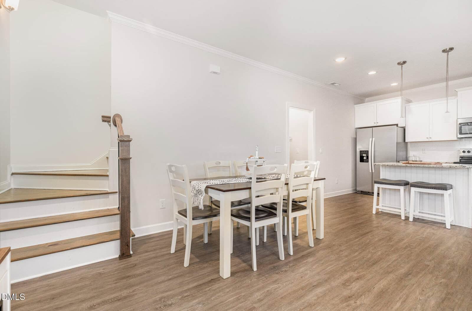 A dining area with a rectangular table and six chairs is next to a kitchen with white cabinets, stainless steel appliances, and a breakfast bar with two stools. A staircase is visible on the left. The floor is light wood.