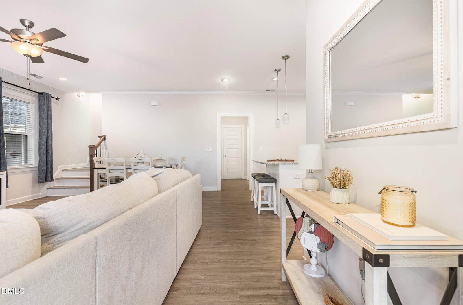 A modern living room with a beige sectional sofa, a ceiling fan, wood flooring, a console table with decor items, and a dining area with a white table and chairs. Stairs lead up to another level.