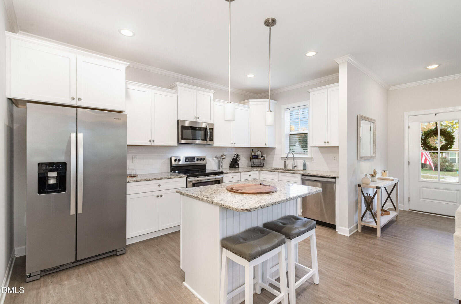 Bright, modern kitchen with white cabinets, stainless steel appliances, an island with two stools, and pendant lighting. There is a window above the sink and a door leading outside. The floor is light-colored wood.