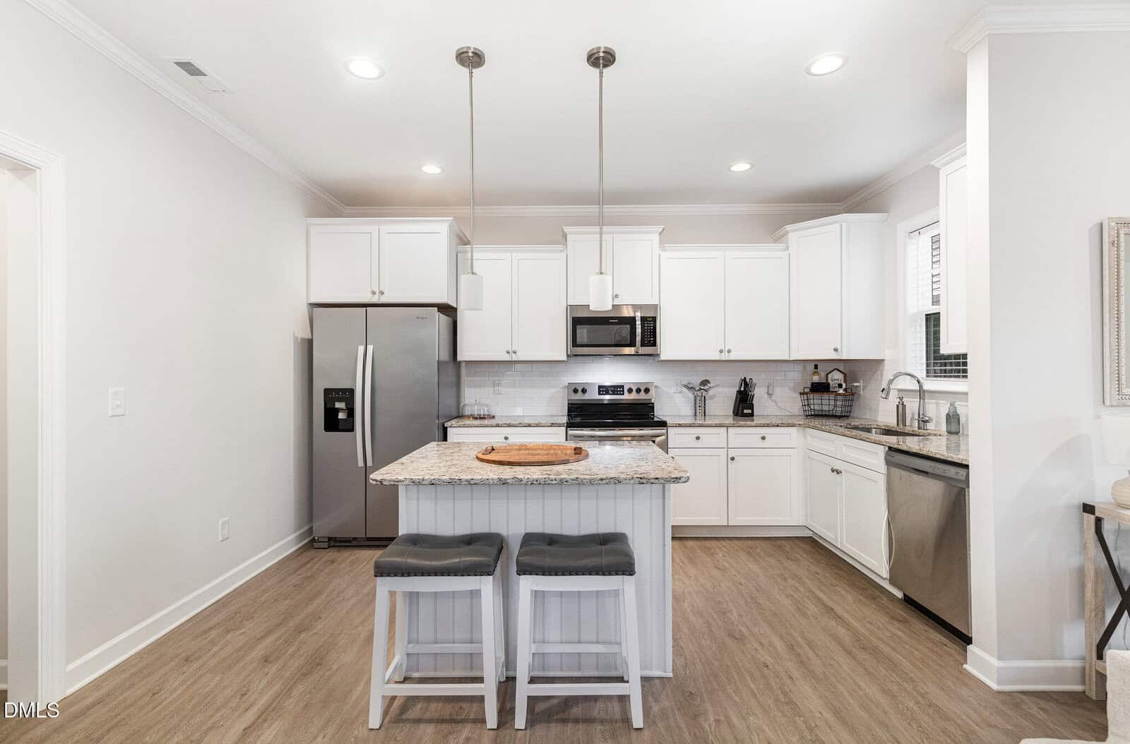 A modern kitchen with white cabinets, stainless steel appliances, a central island with a marble countertop, two gray cushioned stools, wooden flooring, and pendant lights hanging above the island.