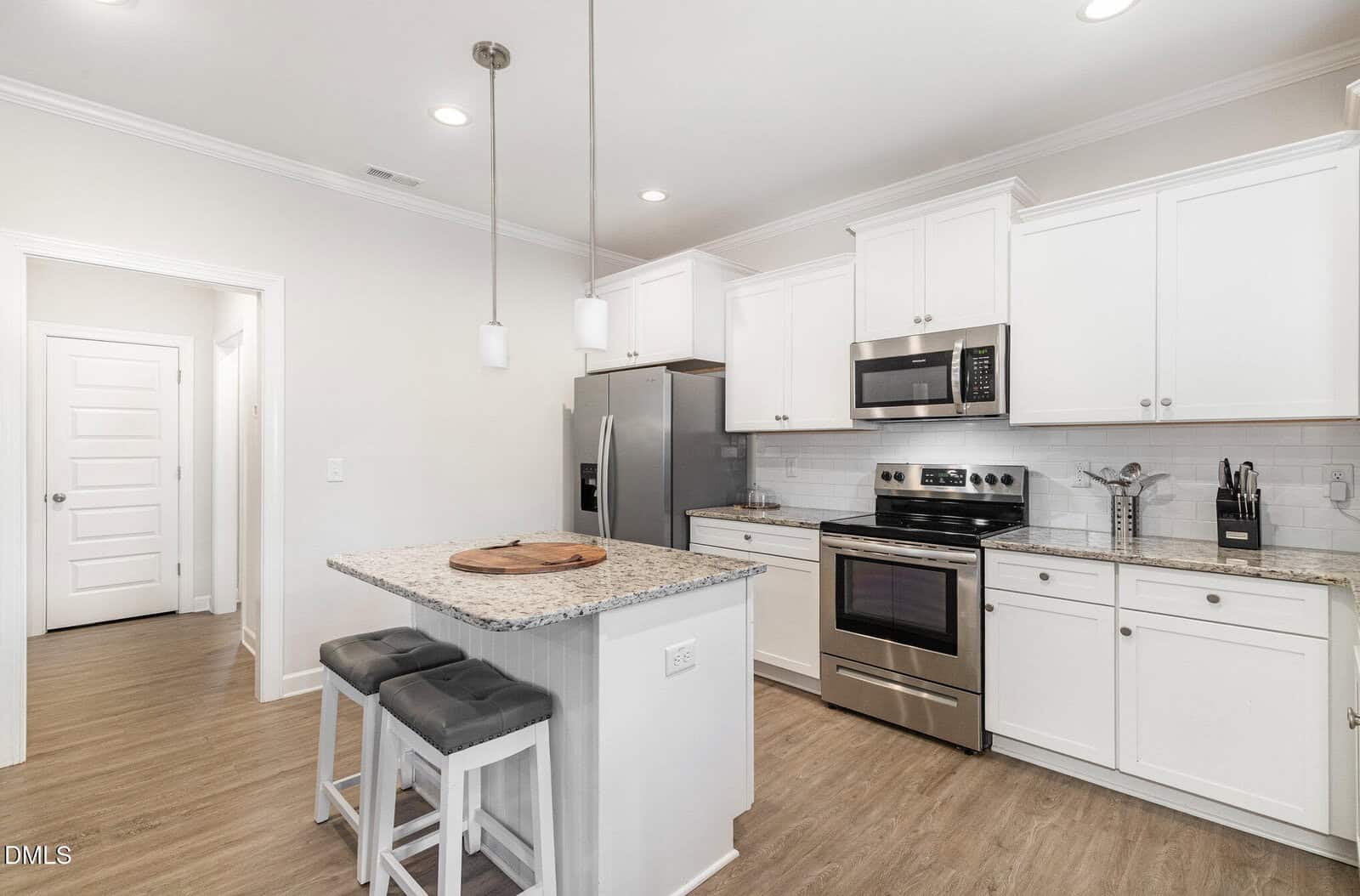 Modern kitchen with white cabinets, stainless steel appliances, granite countertops, a small island with two gray stools, pendant lights, and light wood flooring. A hallway and several doors are visible in the background.