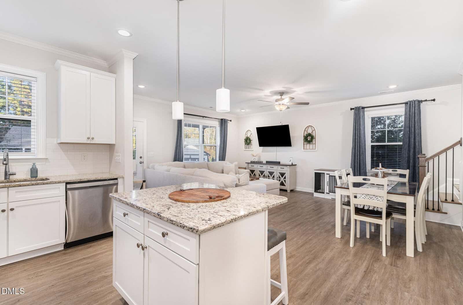 Open-concept kitchen and living area with white cabinets, granite island, stainless steel appliances, dining table, light wood floors, and a living room with a sectional sofa, TV, and large windows with gray curtains.