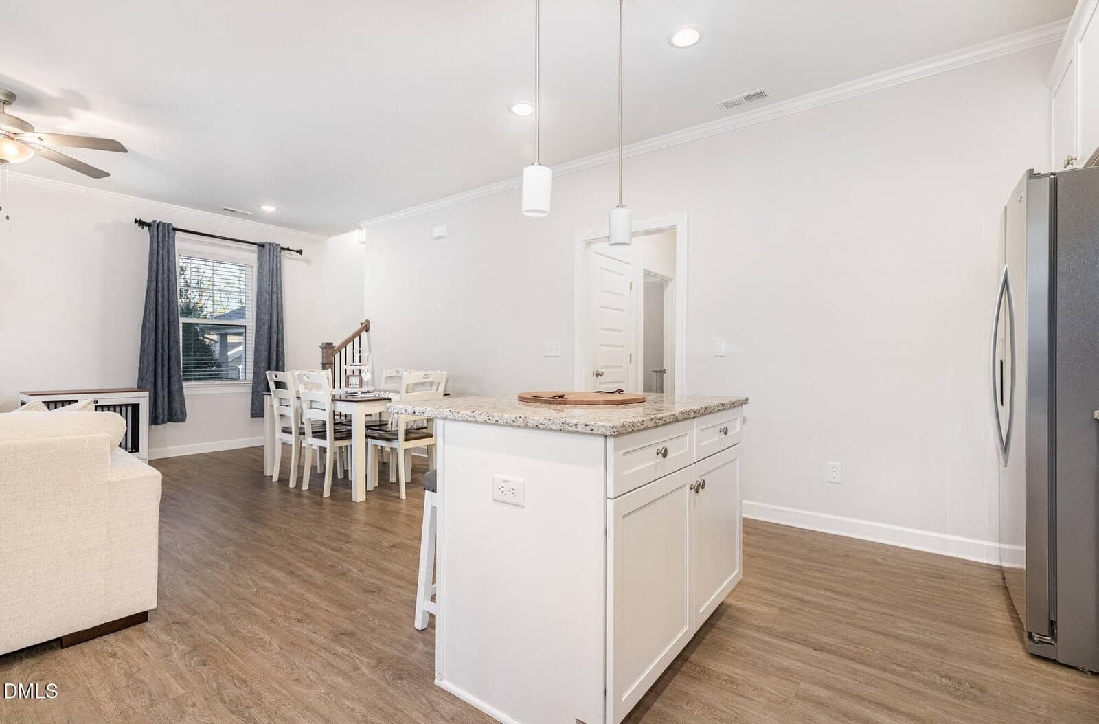 A modern open-concept kitchen and dining area with light wood flooring, white cabinets and countertops, a dining table with chairs, pendant lights, a ceiling fan, and a stainless steel refrigerator.