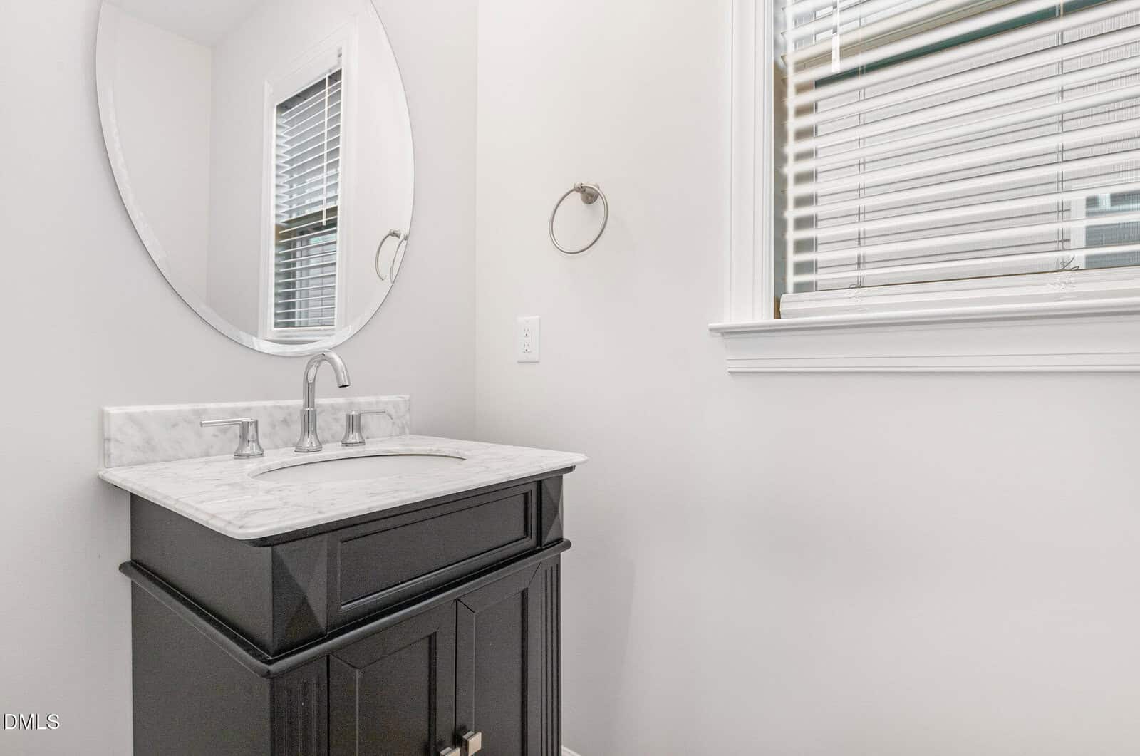 Black bathroom vanity with marble countertop, oval mirror above, wall-mounted towel ring, and a window with blinds. The walls are white, and the overall decor is minimal and modern.