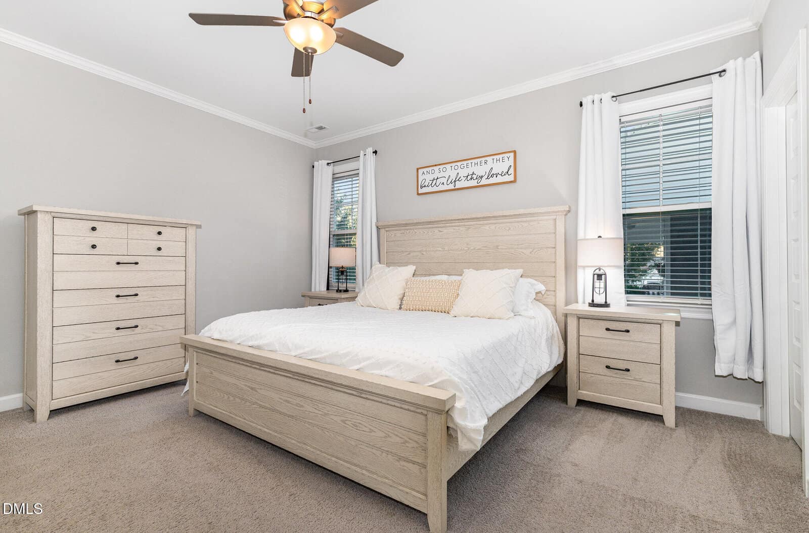 Bright bedroom with light wood furniture, white bedding, two windows with white curtains, a ceiling fan, beige carpet, and a sign above the bed that reads, “So do together they built a life they loved.”.