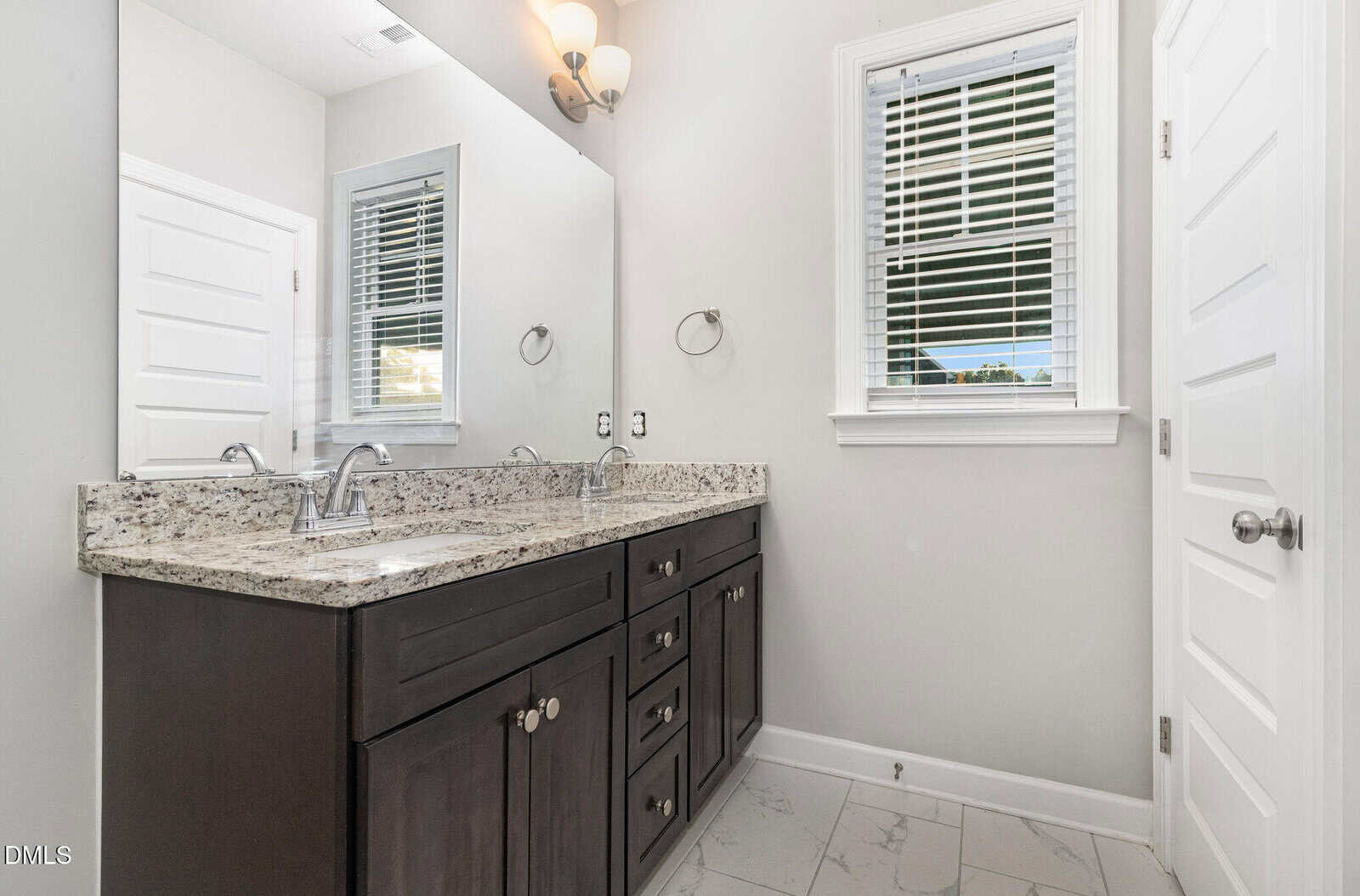 A modern bathroom with a double sink vanity, granite countertop, dark wood cabinets, a large mirror, white walls, tiled floor, window with blinds, and two white doors.