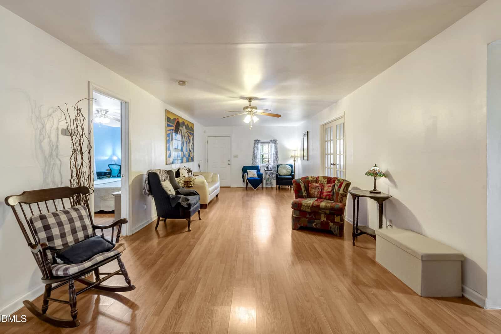 A spacious living room with light wood floors, white walls, several chairs, a small sofa, a ceiling fan with lights, and a side table. A bedroom is visible through an open doorway on the left.
