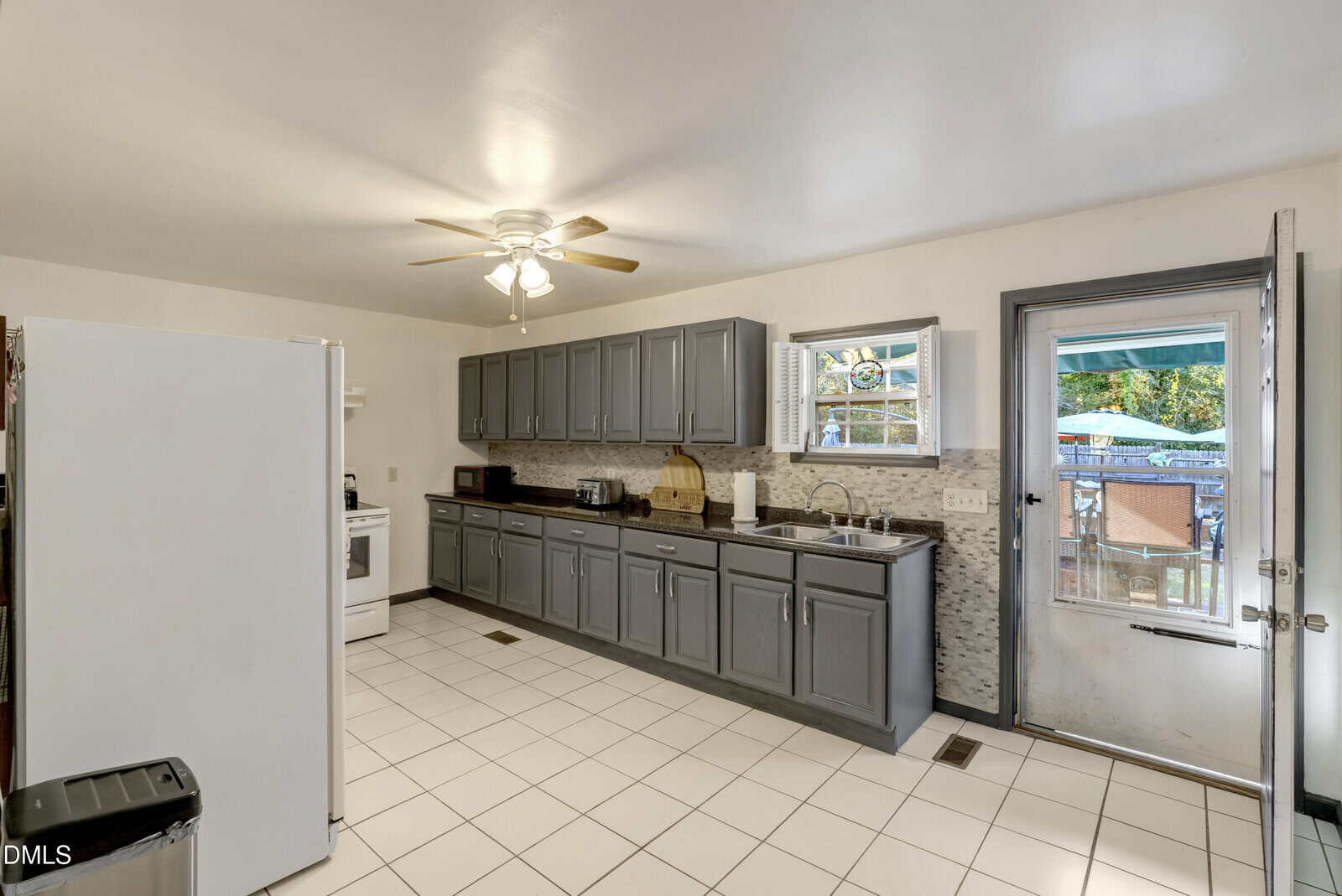 Spacious kitchen with gray cabinets, white appliances, tiled floor, and a ceiling fan. There is a window above the sink and a glass door leading outside to a patio area with outdoor furniture.