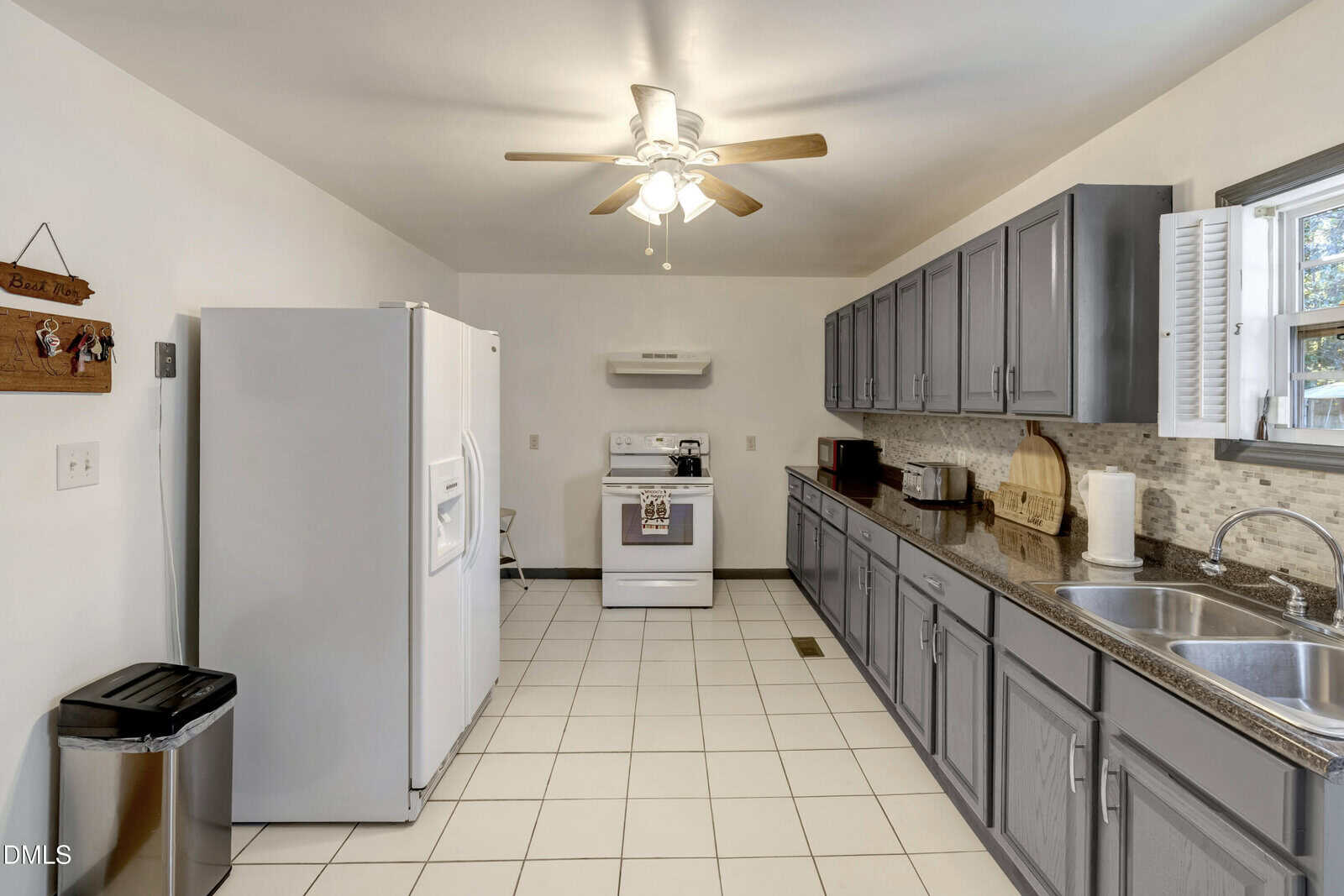 Spacious kitchen with gray cabinets, white appliances, double sink, tile floor, ceiling fan, and a window above the counter.