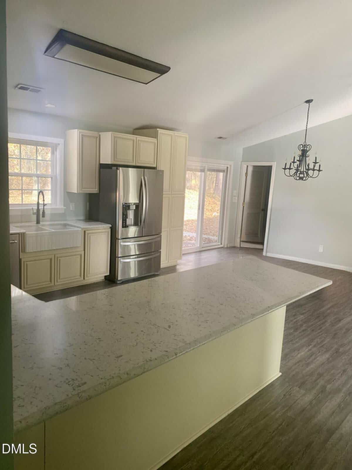 A modern kitchen with white cabinets, a farmhouse sink, stainless steel refrigerator, and light-colored countertops. A chandelier hangs over the adjacent dining area, with glass doors leading outside. Wood floors throughout.