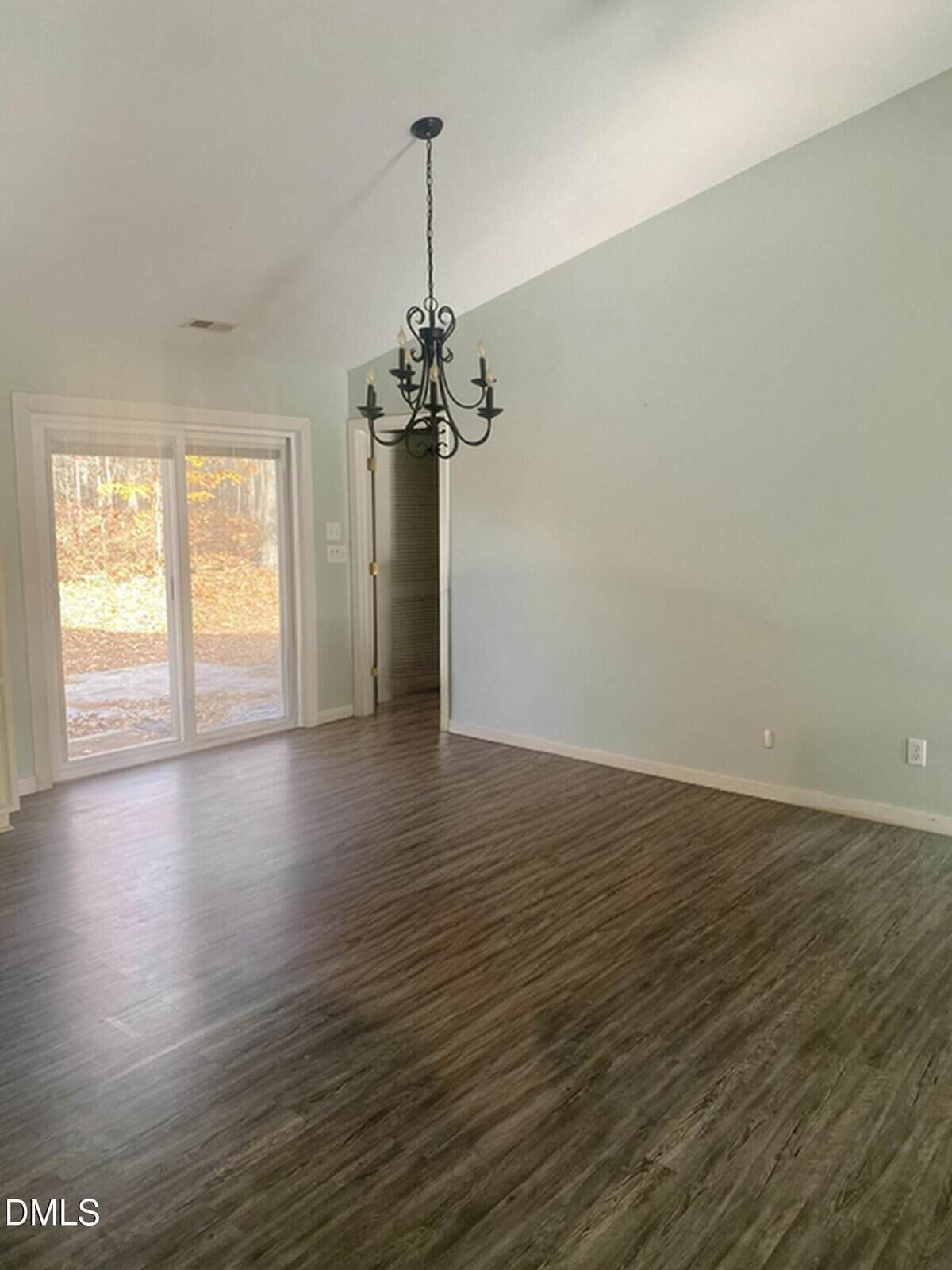 An empty room with light-colored walls, wood-style flooring, a black metal chandelier, sliding glass doors to the outside, and a small doorway leading to another area. Natural light enters from the glass doors.