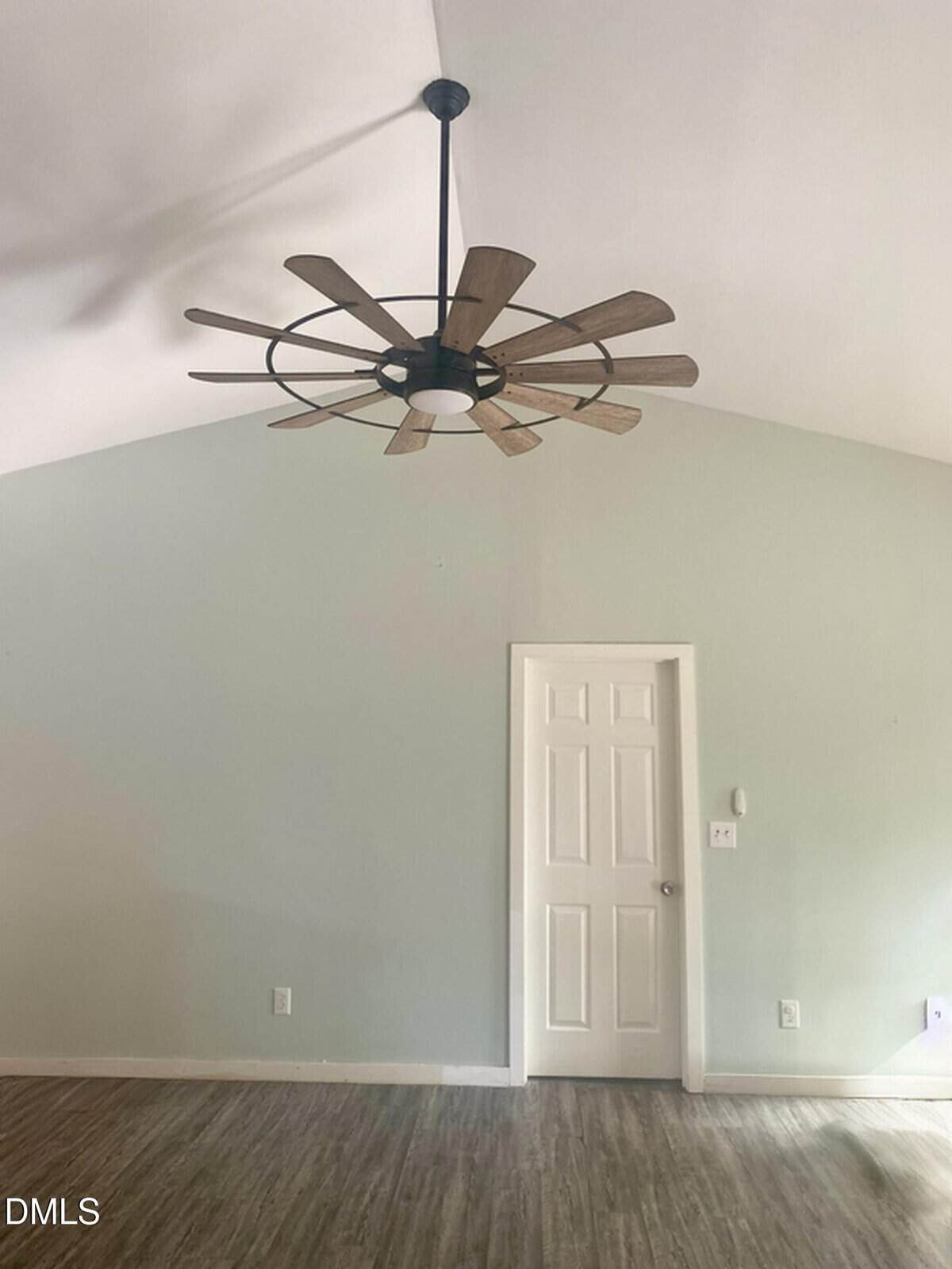A minimalist room with light green walls, a white door, wood-style flooring, and a large ceiling fan with wooden blades. There are three electrical outlets and a light switch visible on the wall.