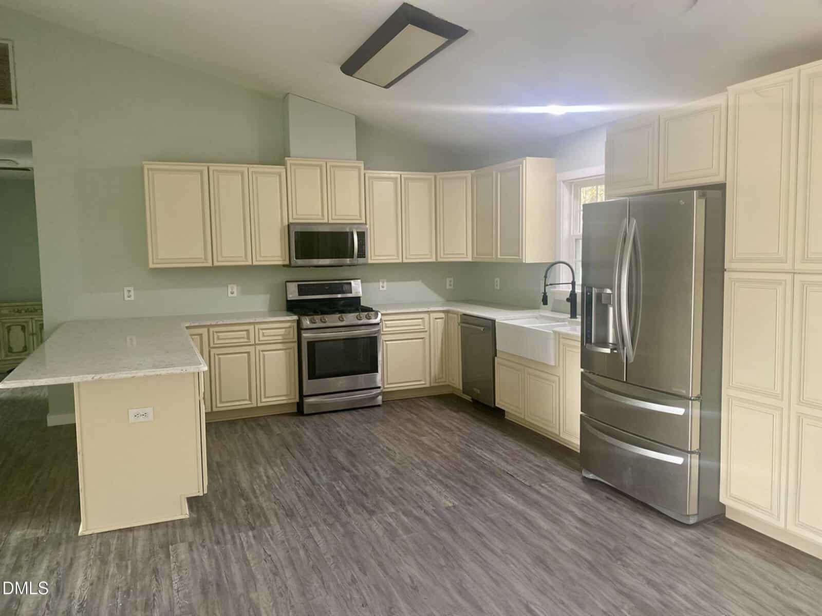 A modern kitchen with cream cabinets, stainless steel appliances including a refrigerator, oven, and dishwasher, a farmhouse sink, and wood-look flooring. A peninsula counter extends from the left side.
