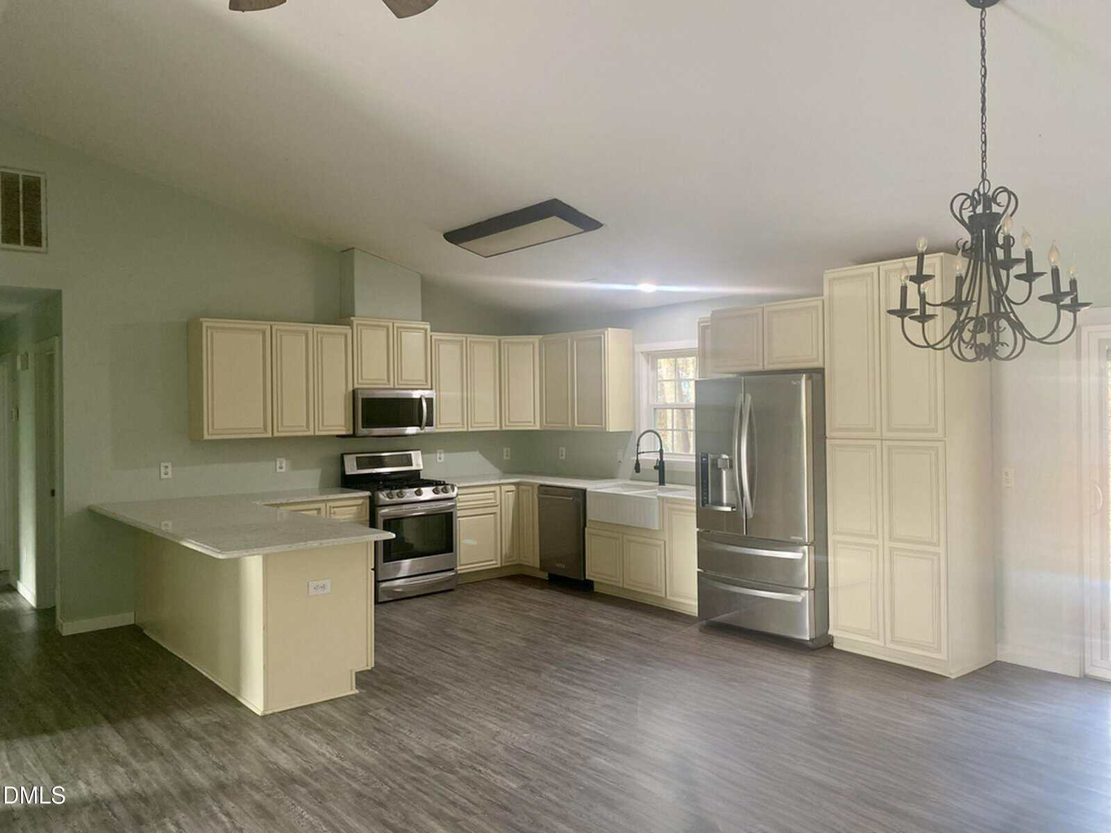 Modern kitchen with cream-colored cabinets, stainless steel appliances, light gray countertops, a large refrigerator, and dark wood-style flooring. A black chandelier hangs near a sliding glass door.