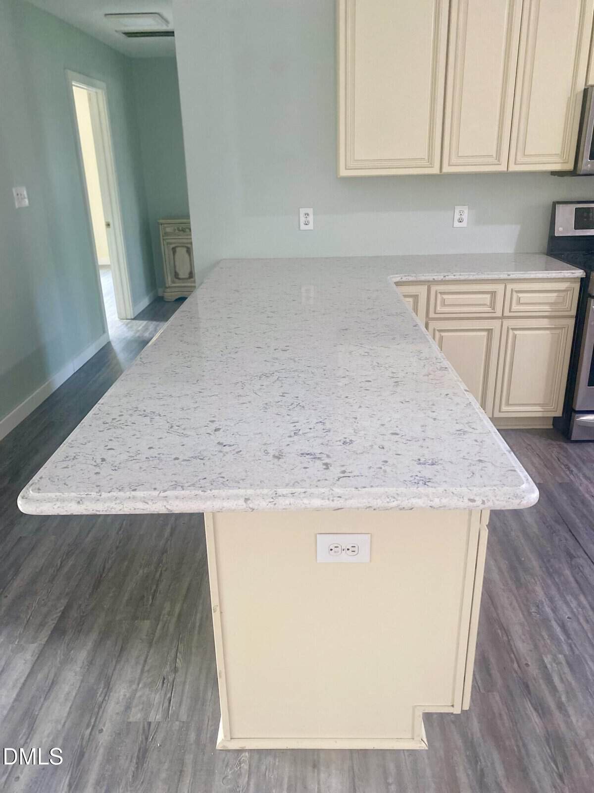 A kitchen with light-colored cabinets and a white granite countertop island. The island has an electrical outlet on the side and gray wood-look flooring underneath. The walls are painted a pale blue-green shade.
