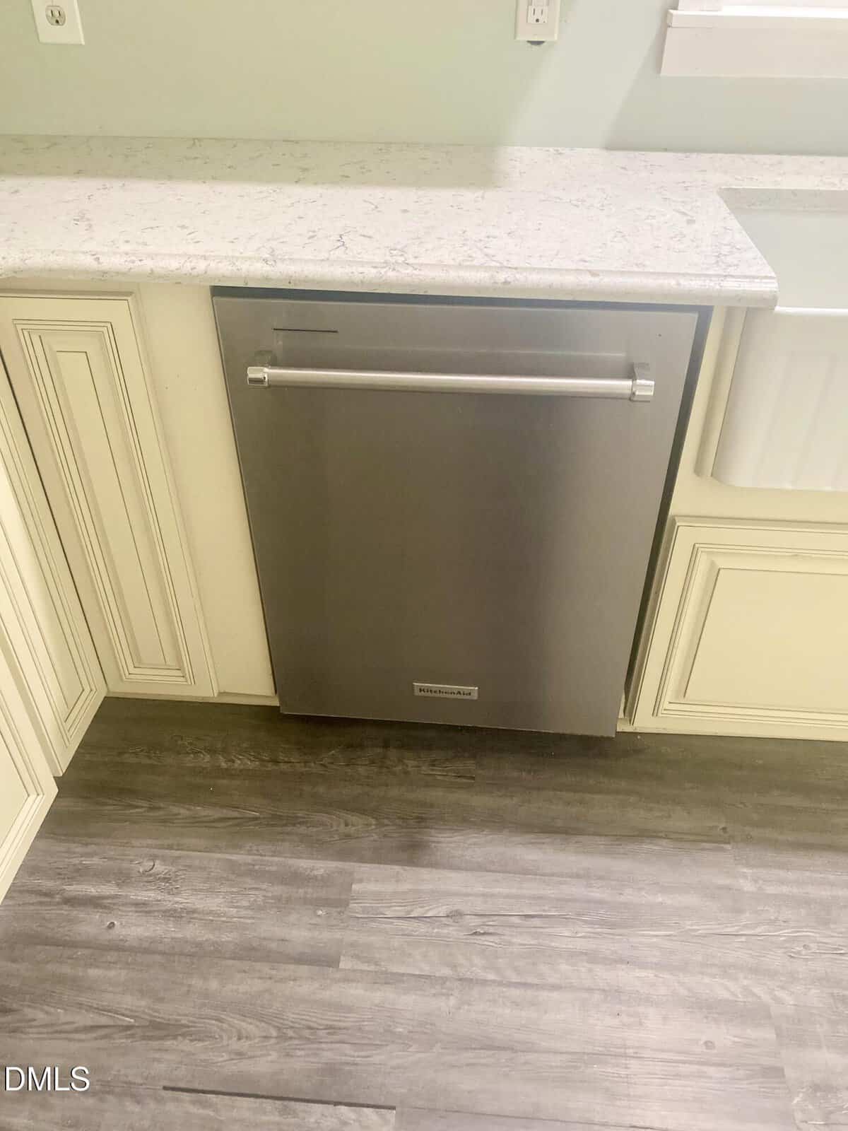 A stainless steel built-in dishwasher is installed under a light-colored marble countertop, surrounded by cream cabinets and gray wood flooring.