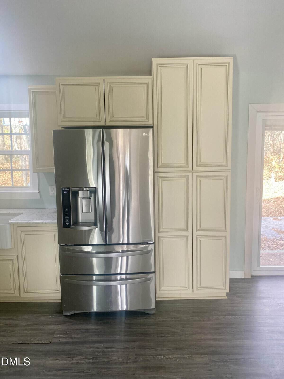 A stainless steel French door refrigerator is surrounded by cream-colored cabinets in a kitchen with light walls and dark wood flooring. A window and glass door let in natural light.