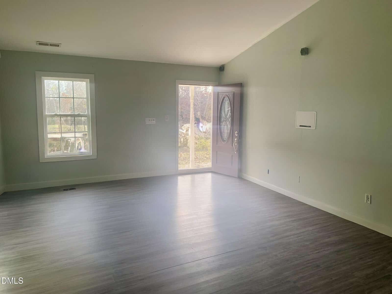 An empty room with light green walls, wood-style flooring, a window on the left, a glass sliding door and a front door in the center, and minimal natural light entering the space.