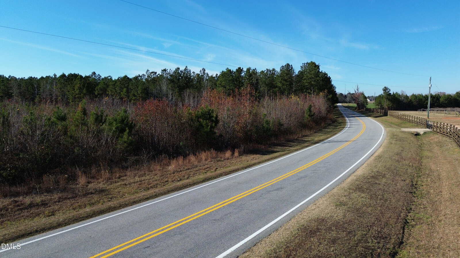 A two-lane rural road curves through a landscape of grass and trees under a clear blue sky, bordered by a wooden fence on one side and a forested area on the other.