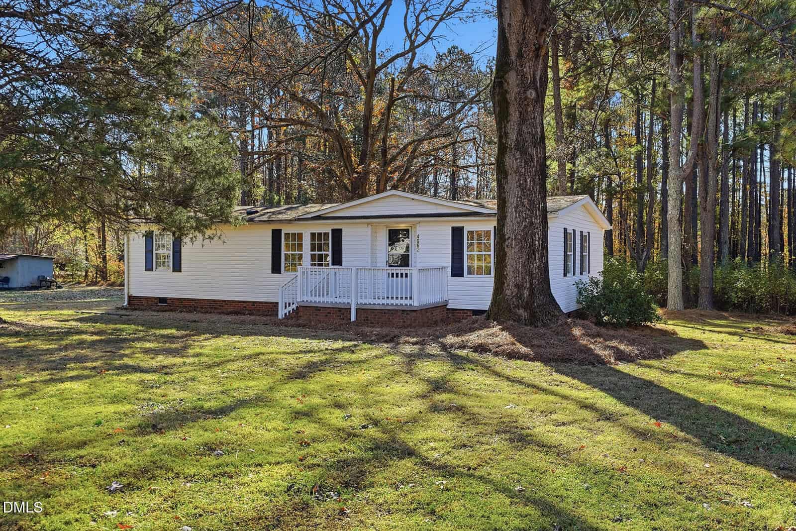 A single-story white house with black shutters and a covered front porch sits on a grassy lawn with large trees and a wooded area in the background.