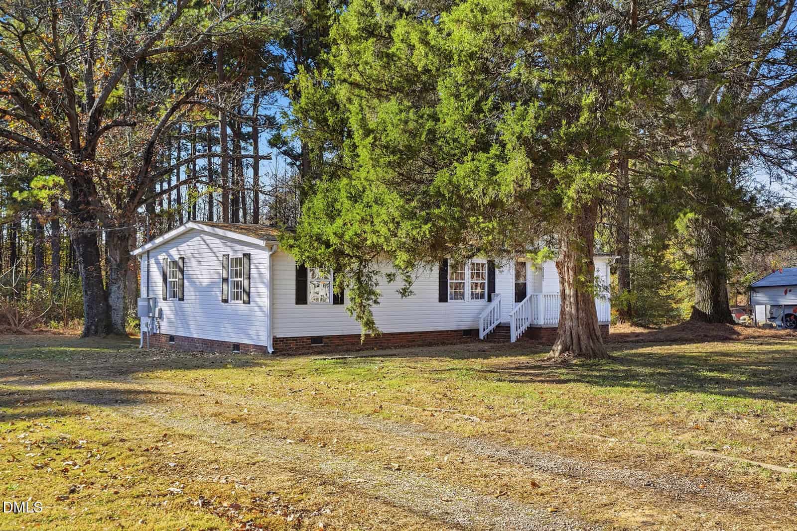 A white single-story house with a small front porch sits on a grassy lot, partially shaded by large trees. The house has brick foundation accents and is surrounded by a wooded area. A driveway is visible in the foreground.