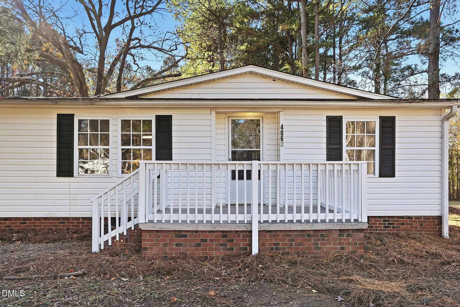 A single-story white house with black shutters, a small covered front porch with white railings, brick foundation, and surrounded by trees. The house number 406 is visible next to the front door.