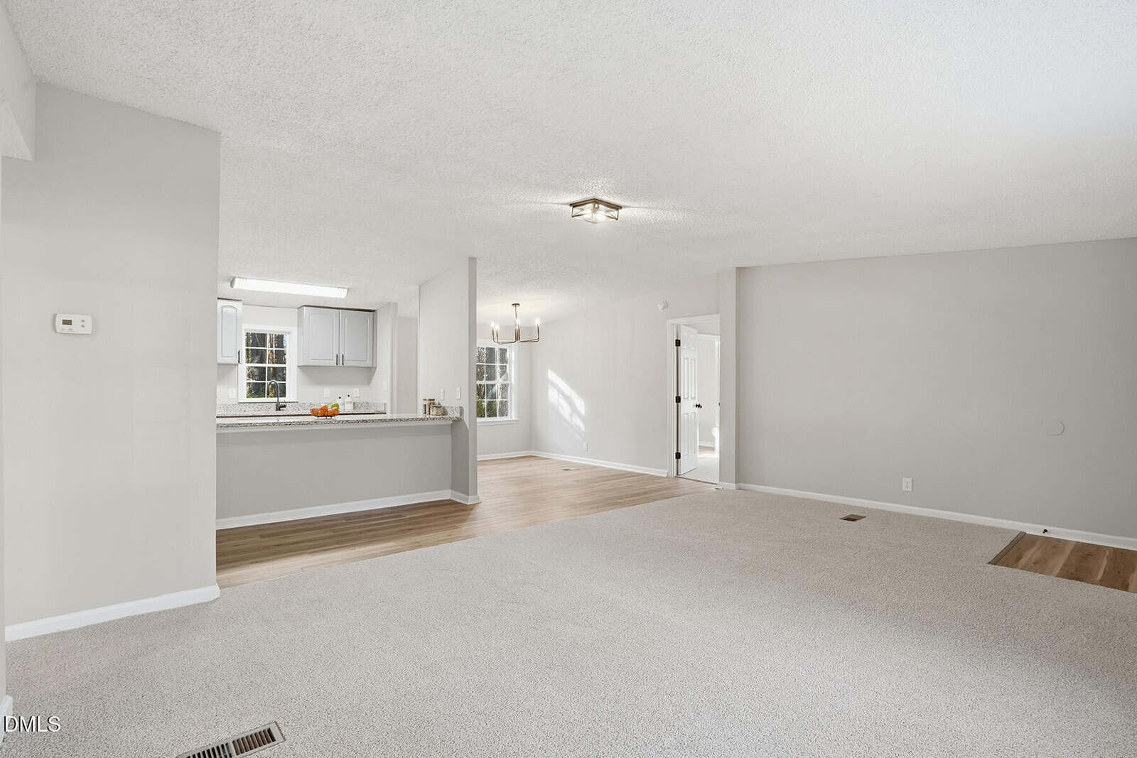 A spacious, unfurnished living and dining area with light gray walls, beige carpet, and wood flooring near the kitchen. The kitchen features white cabinets and a breakfast bar. Large windows provide natural light.