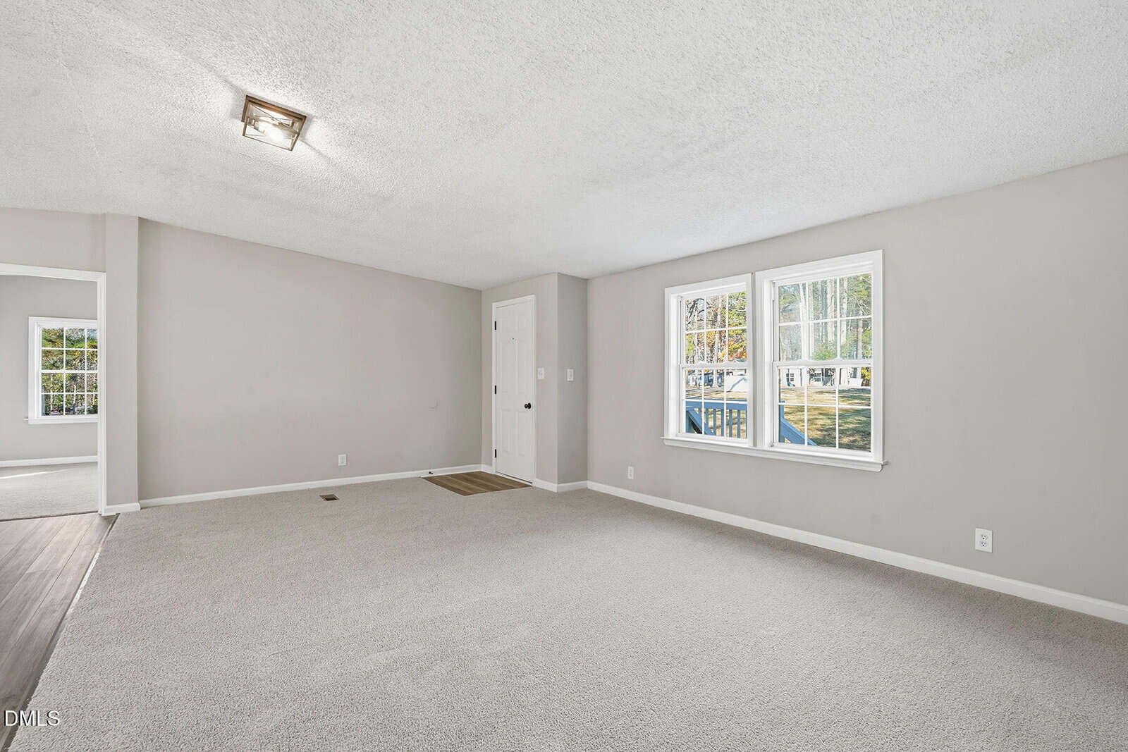 A spacious, empty room with light gray walls, white trim, and beige carpet. Two windows let in natural light. There is a white door and a vent near the corner, and a small area of wood flooring near the door.
